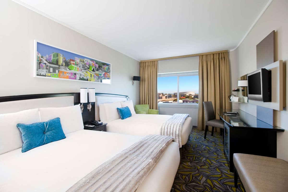 A hotel room with two beds, a desk, a TV, and a large window with a city view.