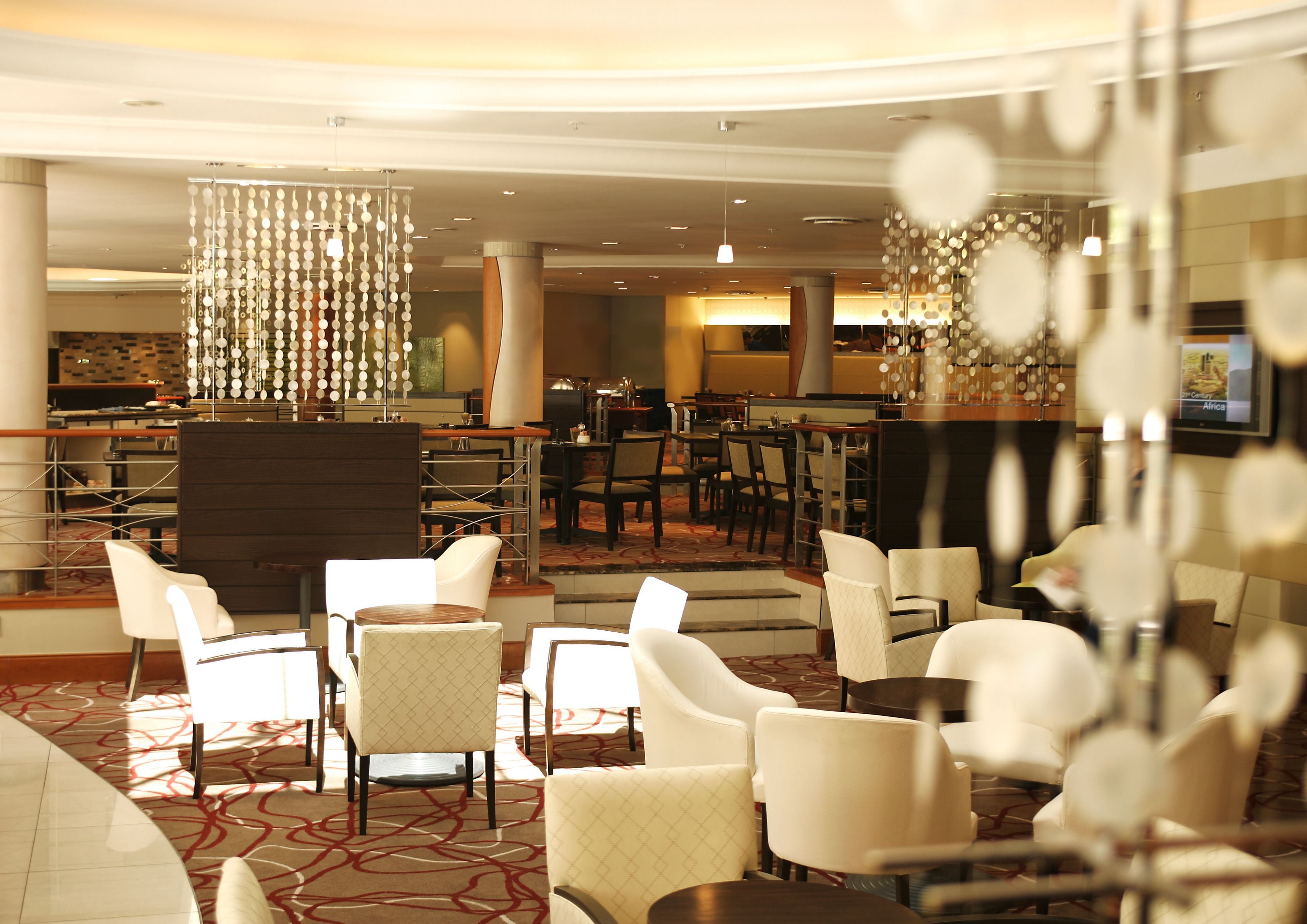 Eine Hotelhalle mit modernem Design, die eine Mischung aus Sitzbereichen und einer Bar bietet. Mehrere Stühle und Tische sind für die Gäste bereitgestellt, und ein großer Bildschirm zeigt Inhalte.