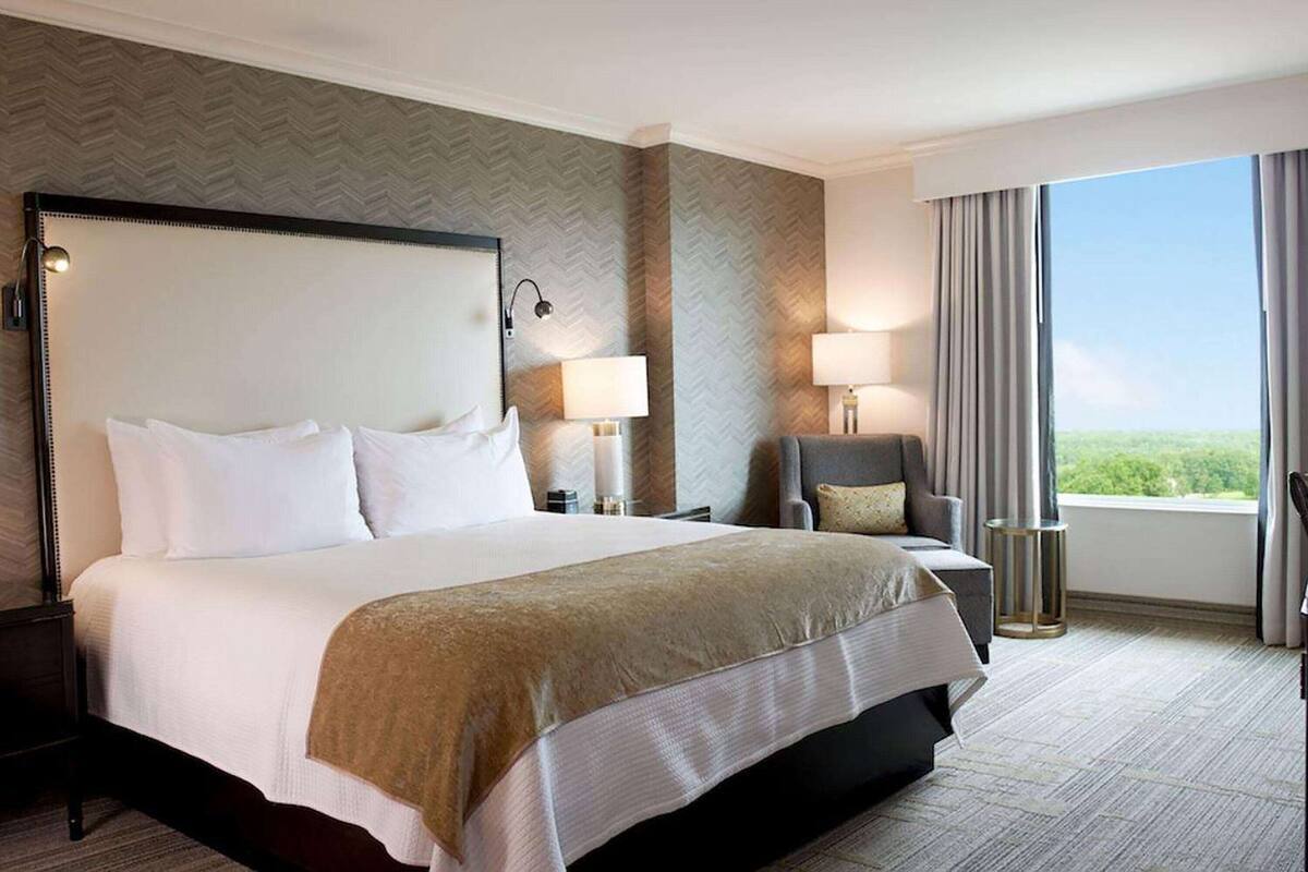 A hotel room with a large bed, two bedside tables with lamps, a chair, and a view of the outdoors.