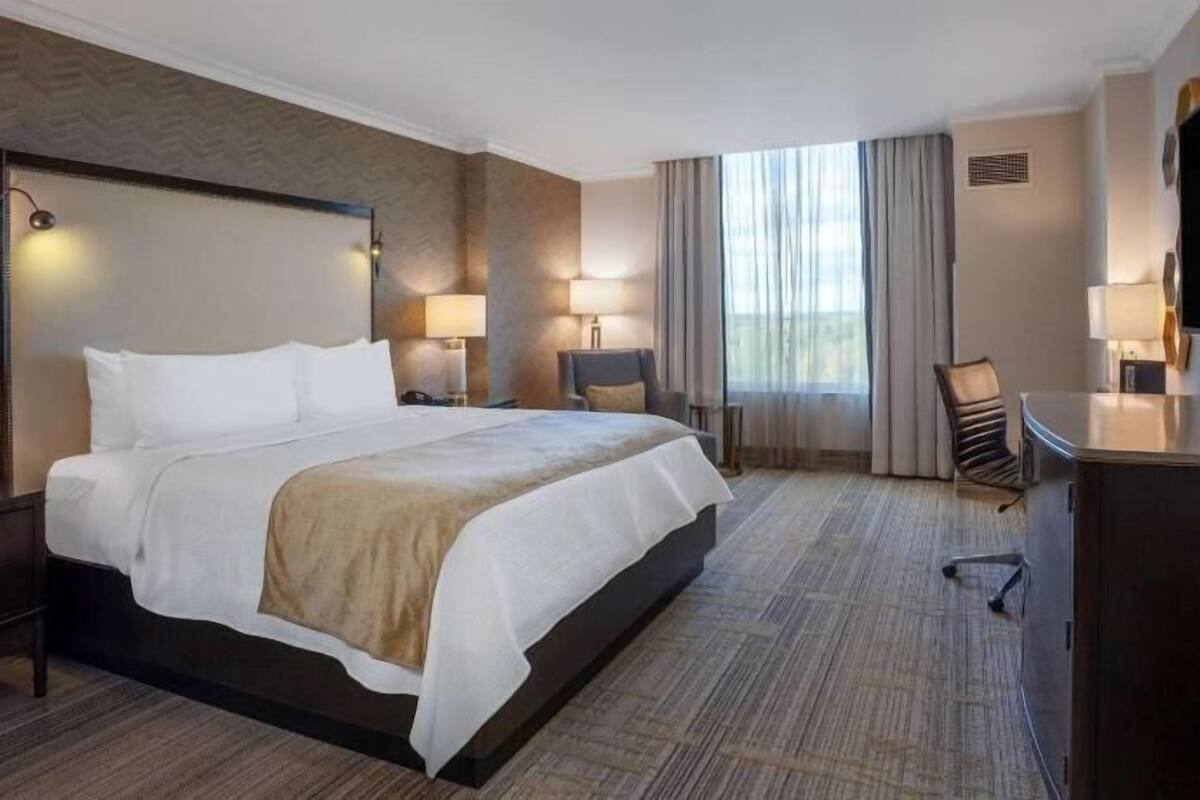 A hotel room with a large bed, two bedside tables, a desk, a chair, a TV, and a window with curtains.