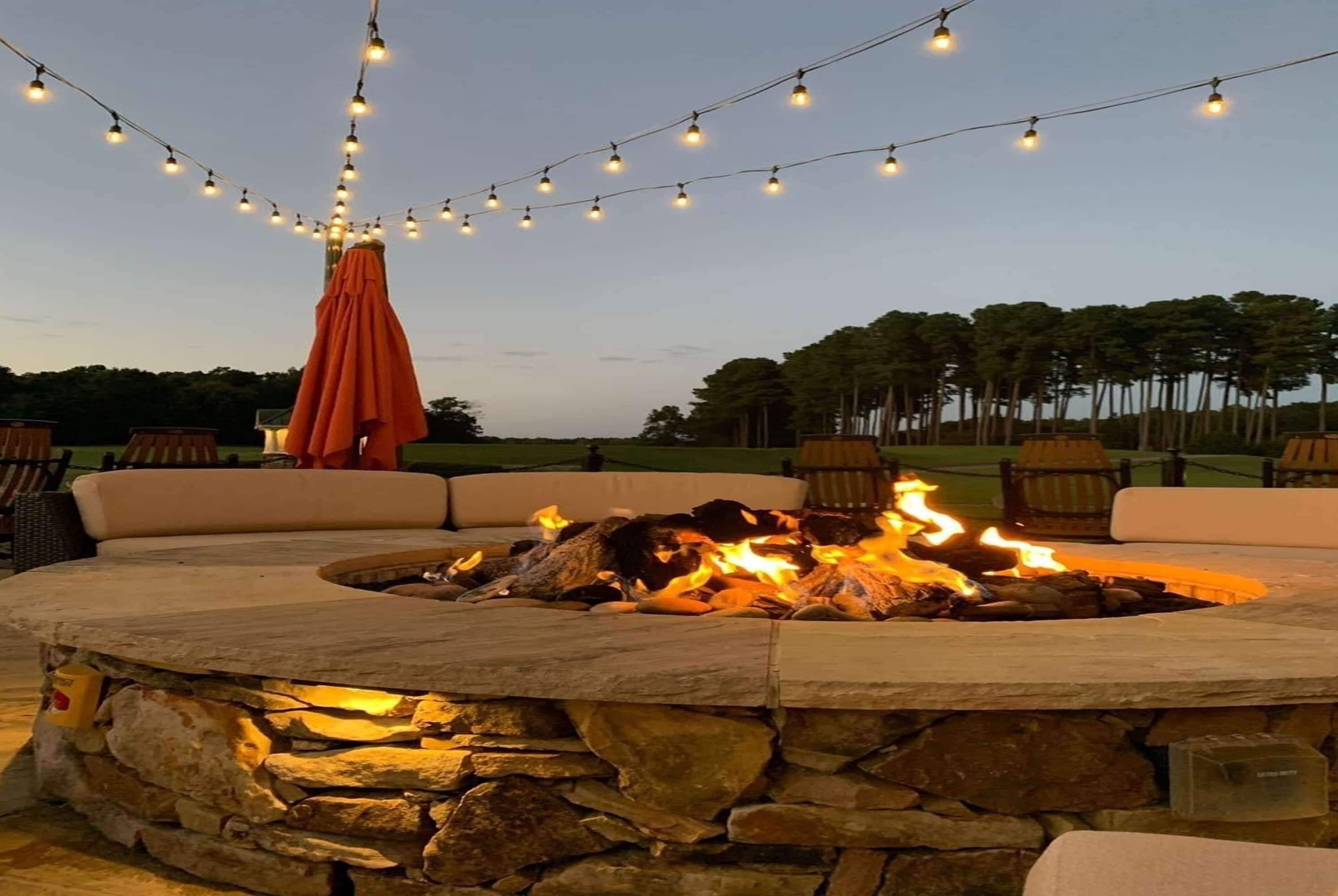 An outdoor seating area with a stone fire pit, string lights, and an orange umbrella.