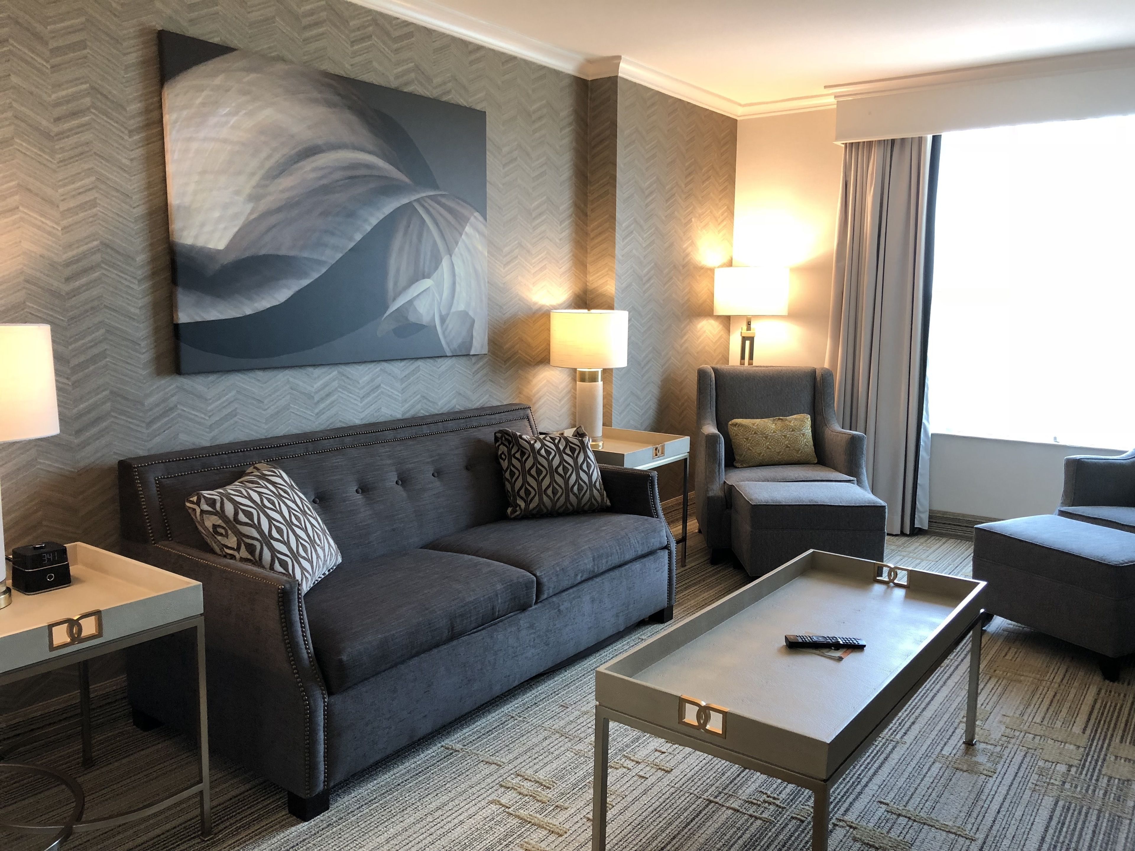 Ein modernes Hotelzimmer mit grauem Sofa, passendem Sessel, einem kleinen Tisch und einem großen, abstrakten Gemälde an der Wand.