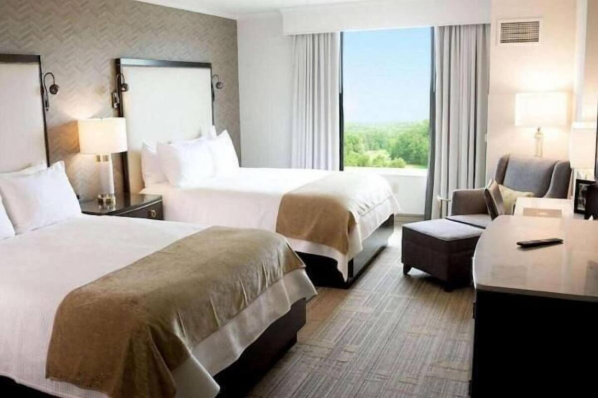 A hotel room with two beds, a desk, a chair, and a large window with a view of greenery.