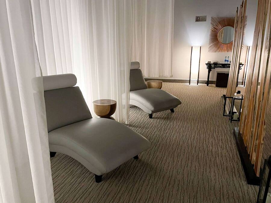 A hotel room with two grey lounge chairs, a wooden side table, a small round table, and a mirror on the wall.