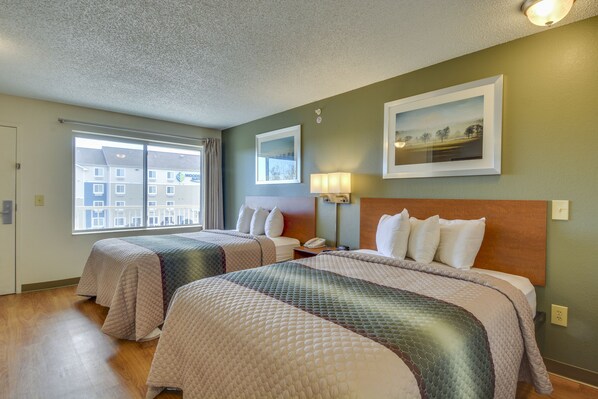 A hotel room with two beds, a large window, a painting on the wall, and a lamp.