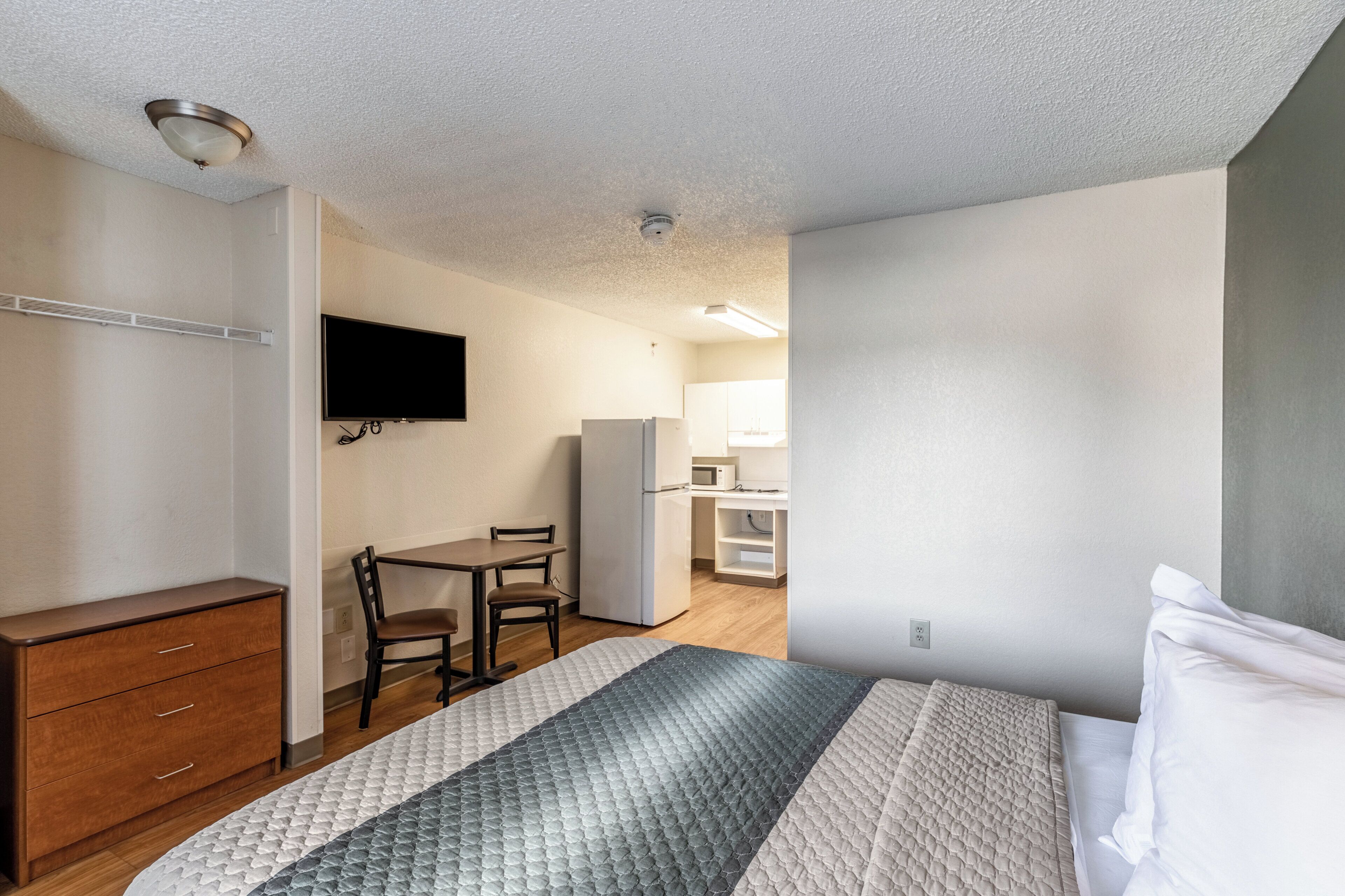 A bedroom with a bed, a desk, a chair, a television, and a kitchenette.