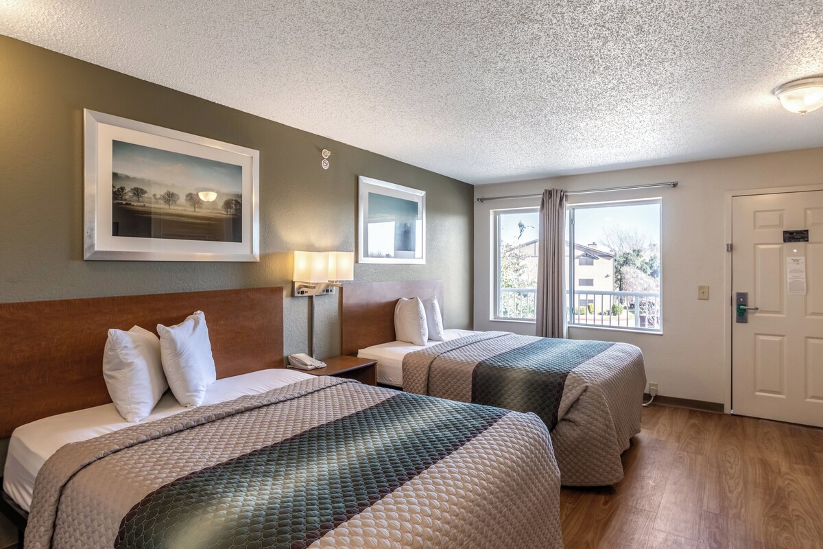 A hotel room with two beds, a nightstand, a lamp, a framed picture on the wall, and a window with curtains.