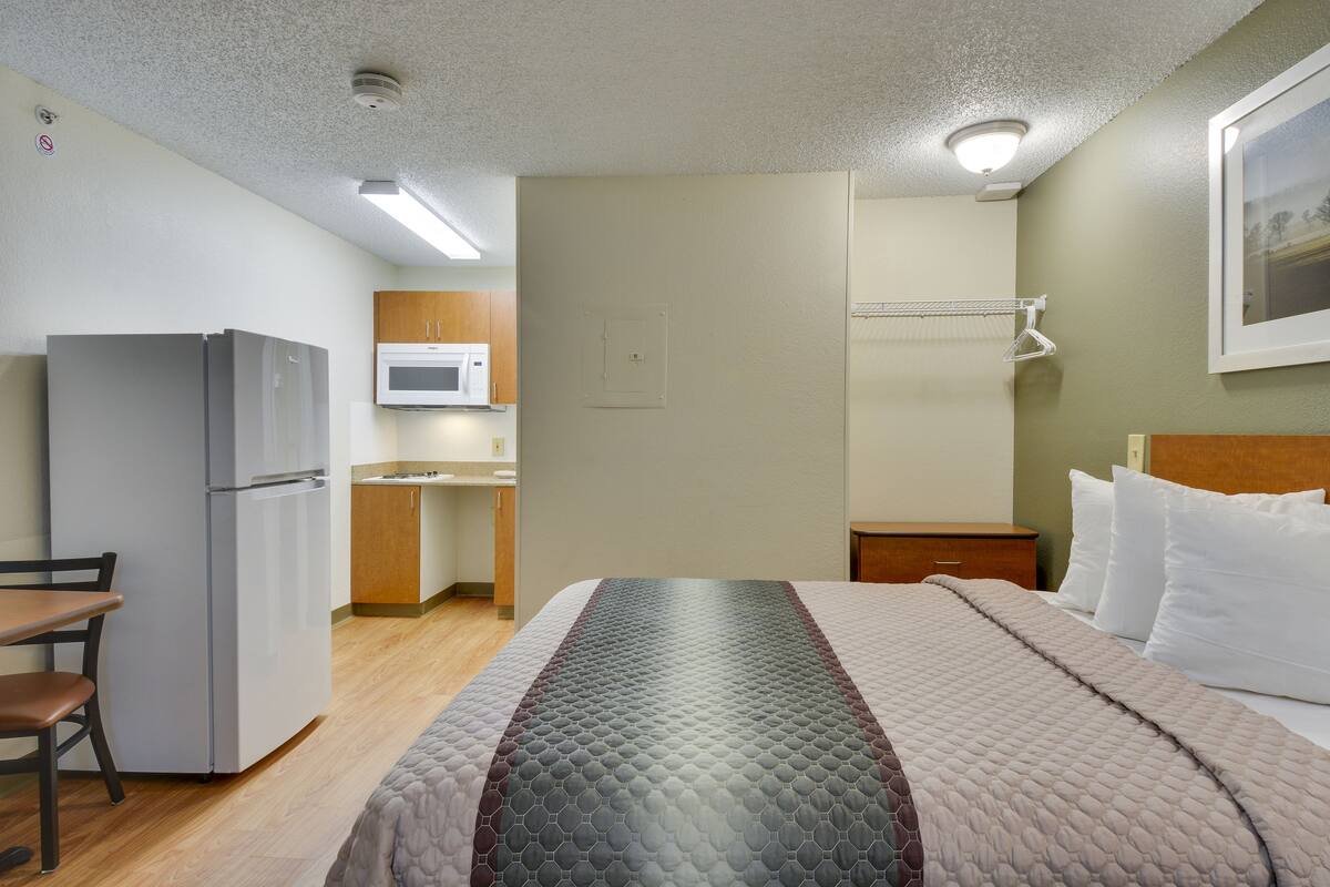 A compact hotel room with a kitchenette, a bed with a quilted bedspread, a wooden nightstand, and a wall-mounted TV.