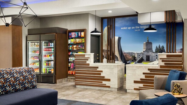 A modern lobby with a seating area, a refreshment bar, and a large poster of a cityscape. - La Quinta Inn & Suites by Wyndham Salem OR (Salem)