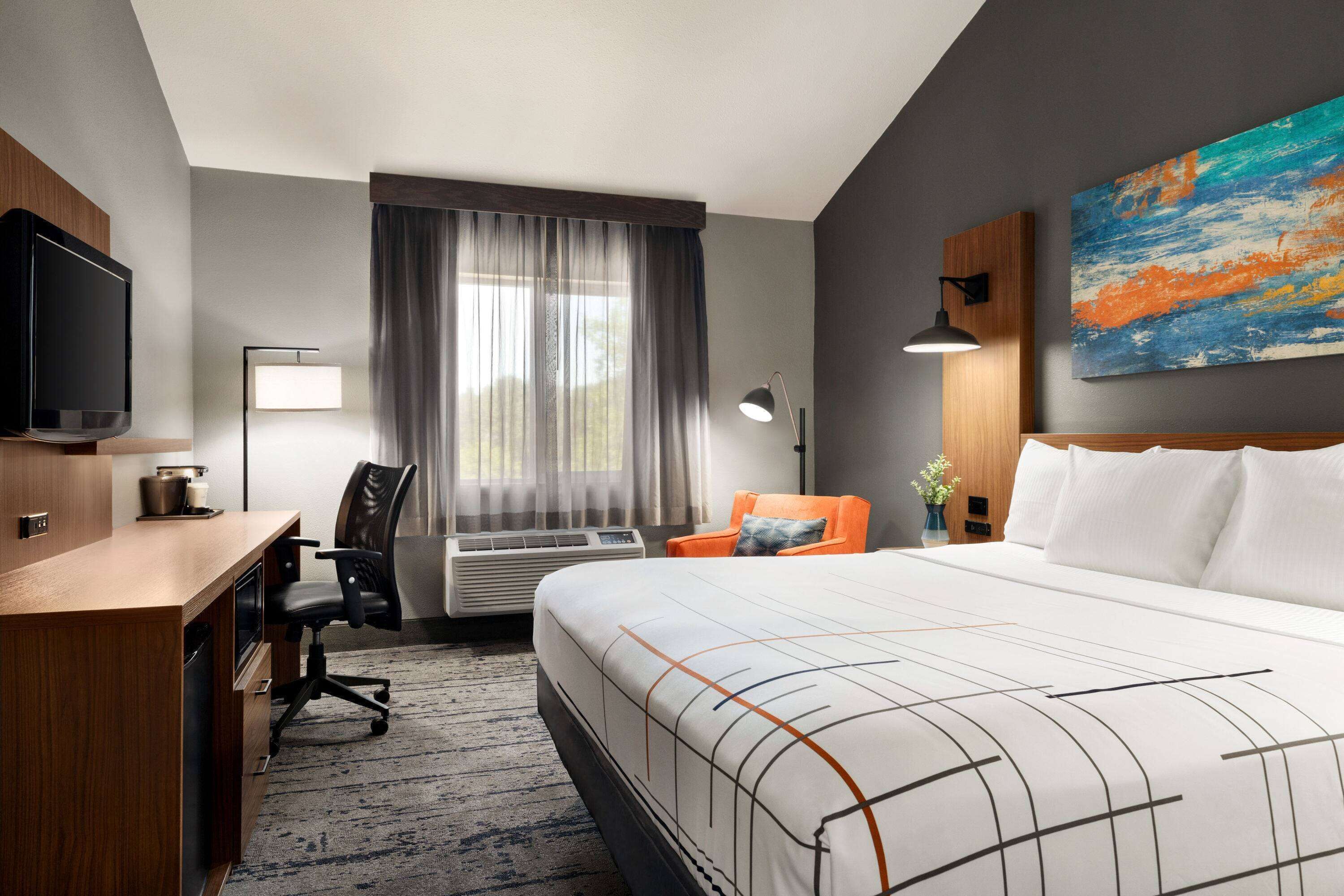 A hotel room with a bed, desk, chair, TV, and artwork on the wall.