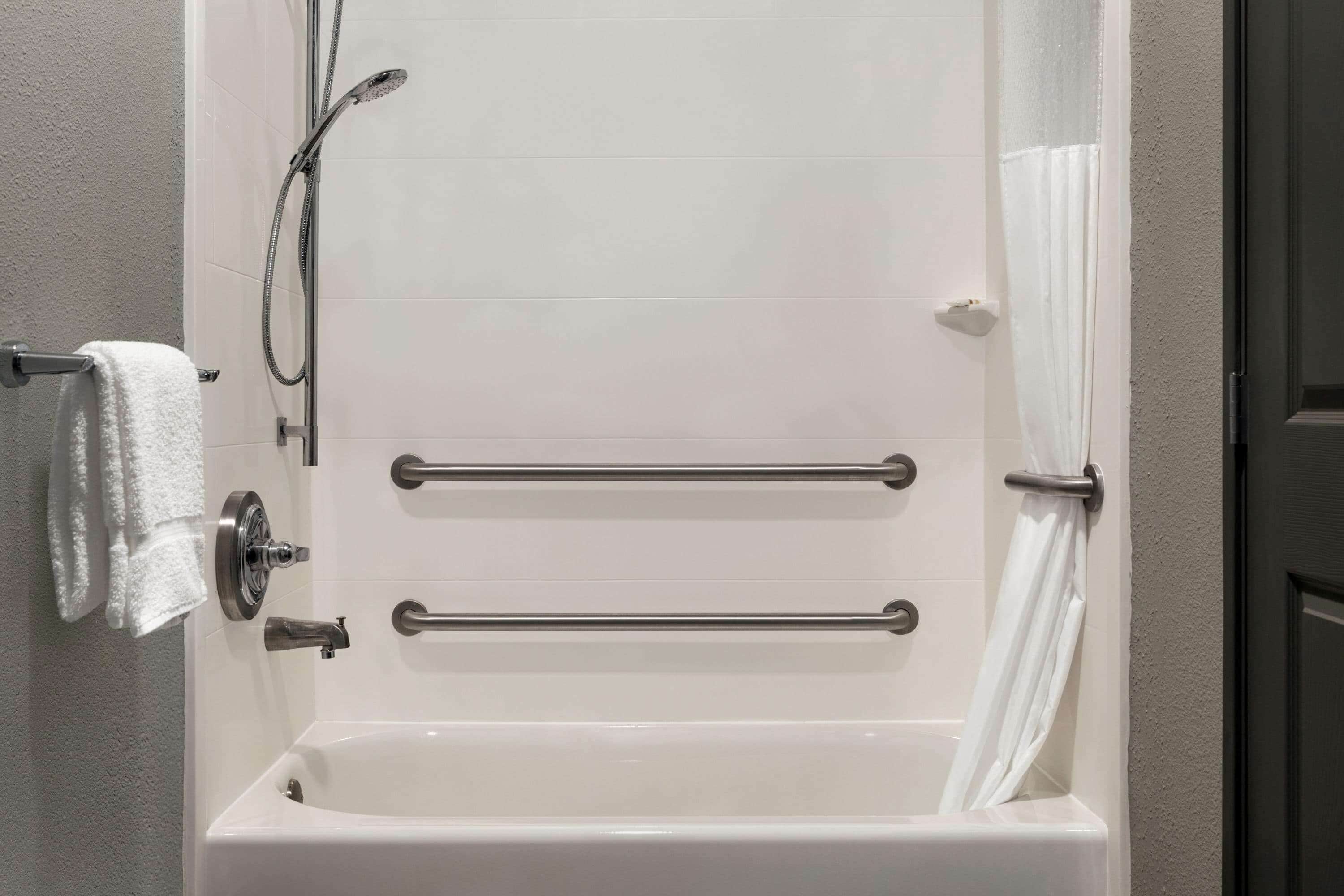 Una bañera blanca con dos barras de apoyo, un cabezal de ducha y un perchero con una toalla.