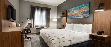 A hotel room with a large bed, a desk, a TV, a painting, and a window with curtains.