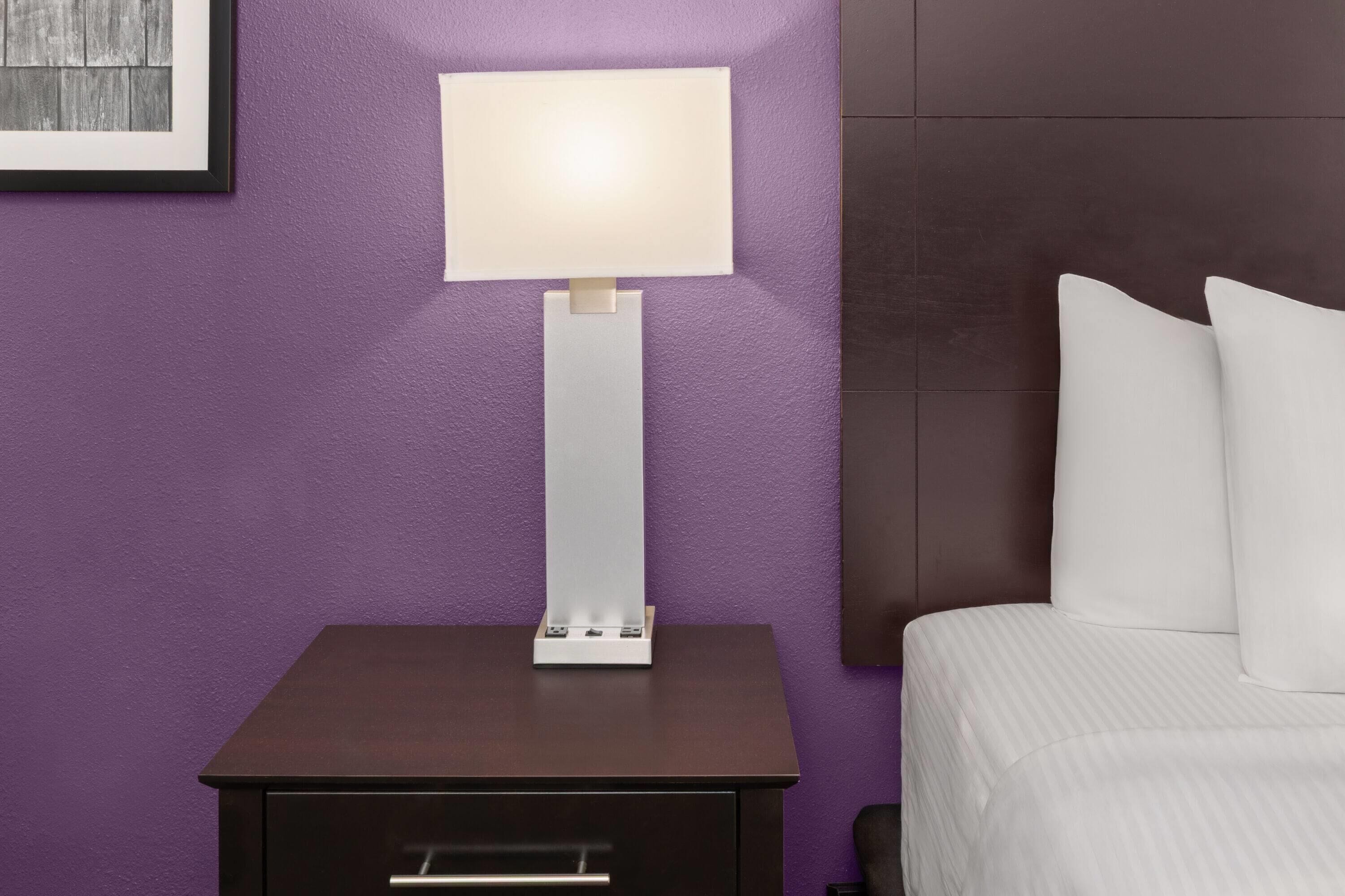 A neatly made bed with white linens, a bedside table with a lamp, and a framed picture on the wall.