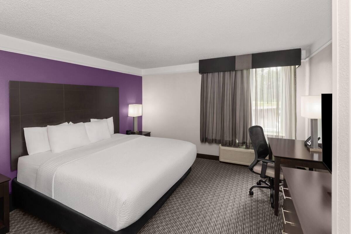 A hotel room with a large bed, a desk, a chair, a lamp, and a window with curtains.