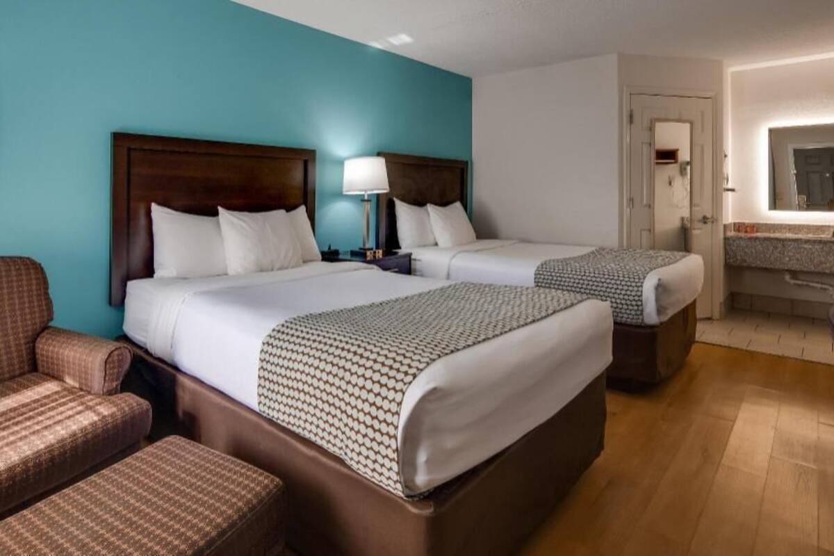 Een hotelkamer met twee bedden, een bank, een nachtkastje, een lamp, een badkamer met een spiegel en een houten vloer.