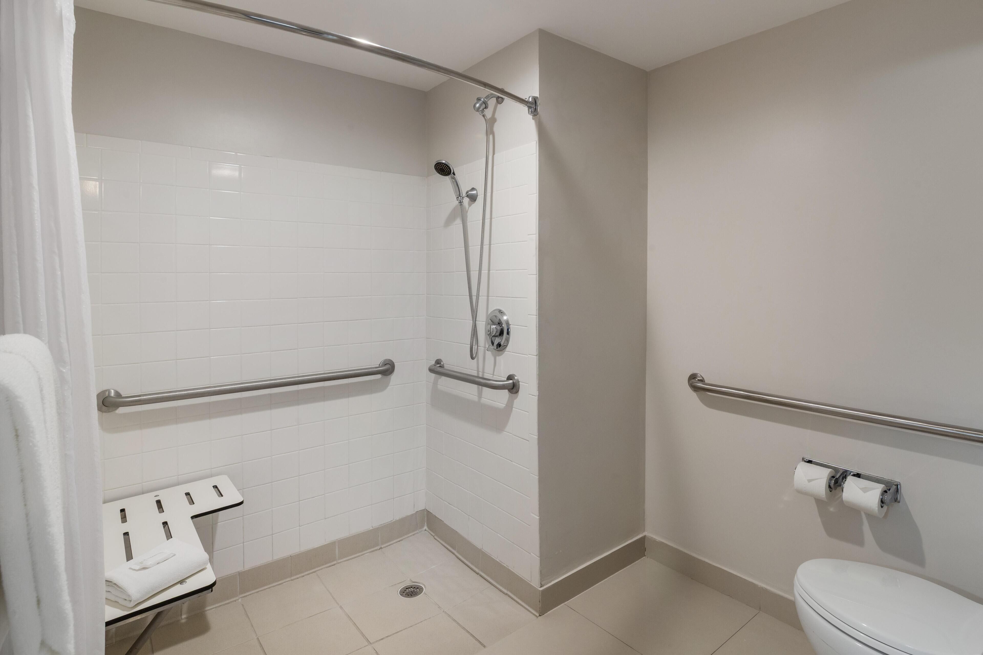 Un baño pequeño y limpio con azulejos blancos, un inodoro, una ducha con barras de apoyo y un perchero para toallas.