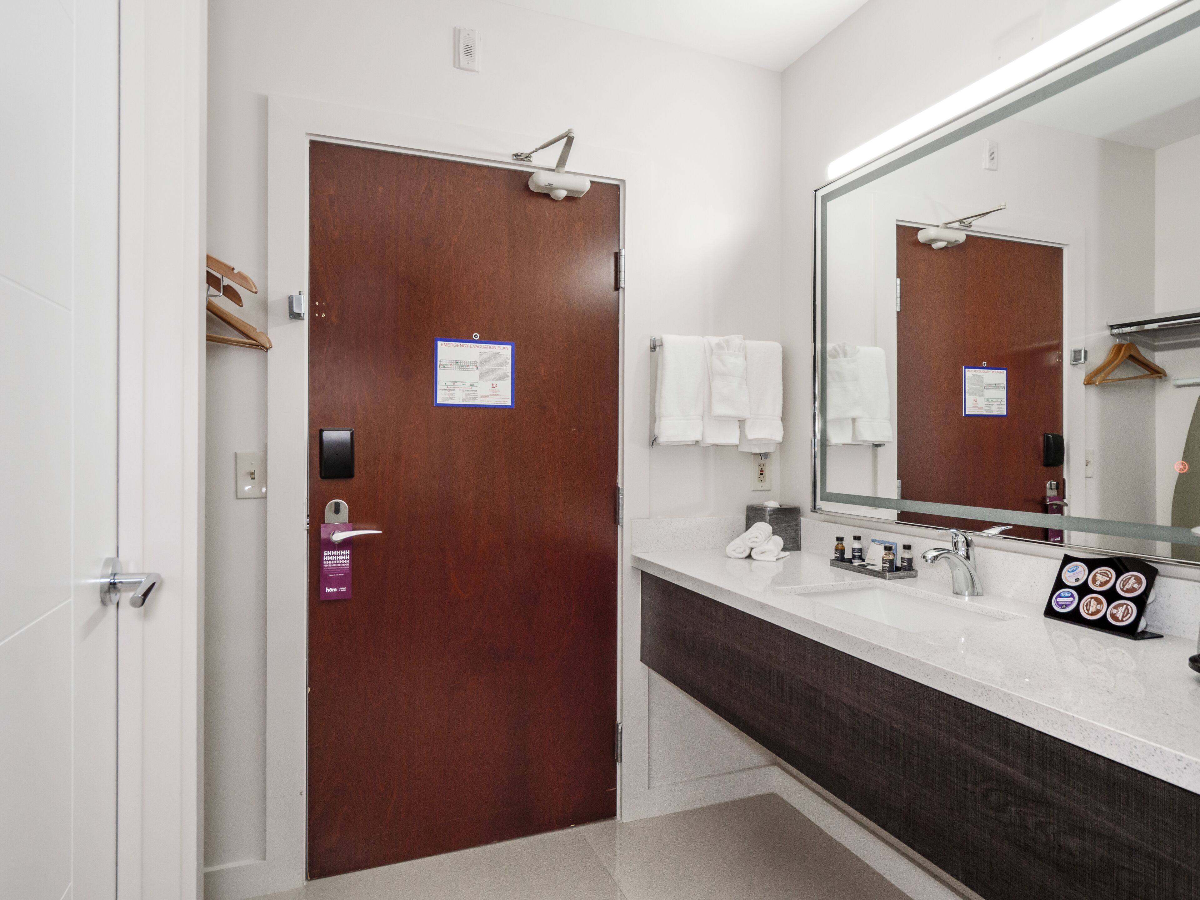 Un baño de hotel con una puerta de madera, un espejo grande, un lavamanos blanco y un perchero para toallas.