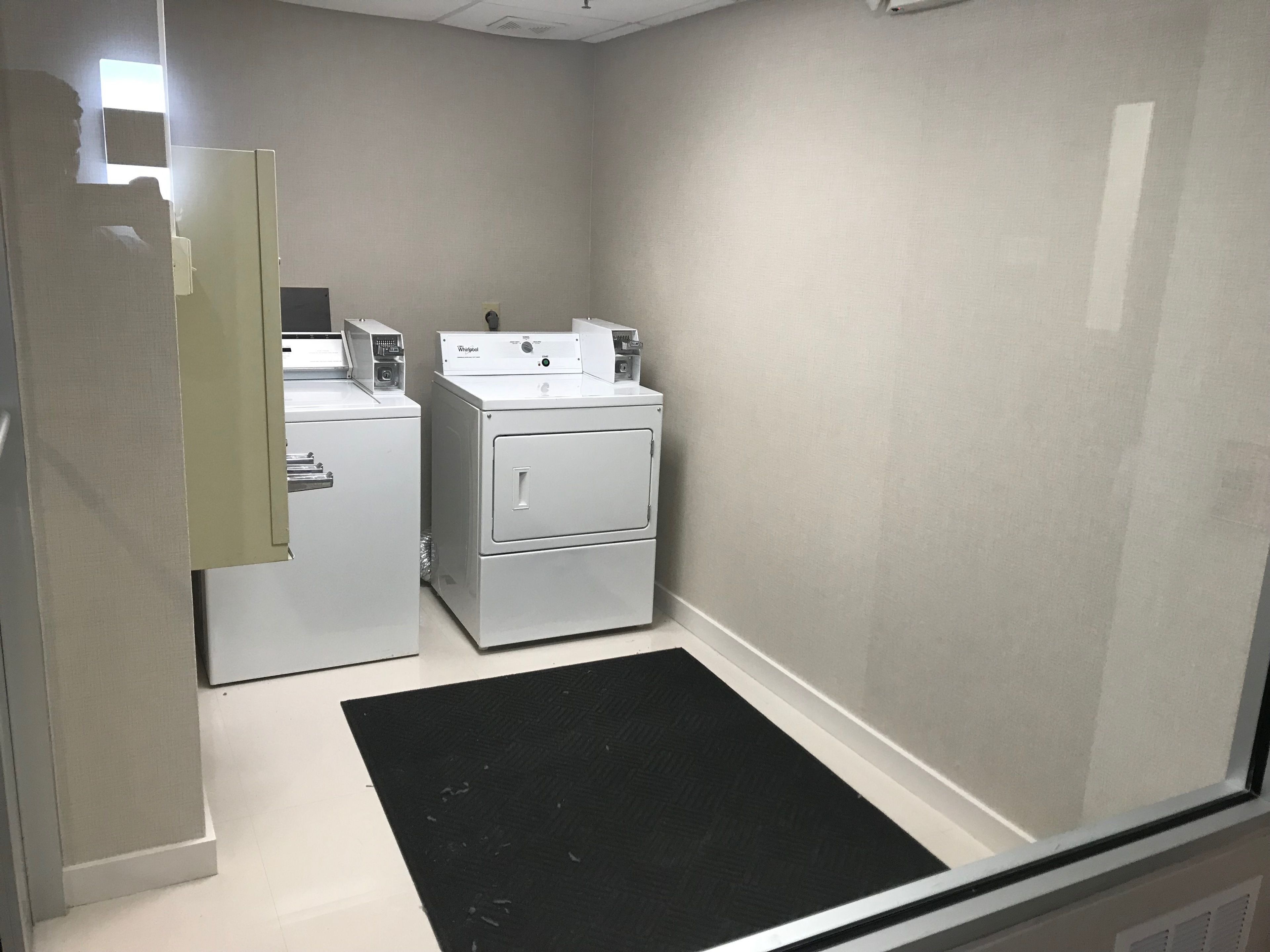 Une buanderie équipée de deux lave-linge et deux sèche-linge Whirlpool, d’un tapis noir à l’entrée et d’une porte donnant sur une autre pièce.