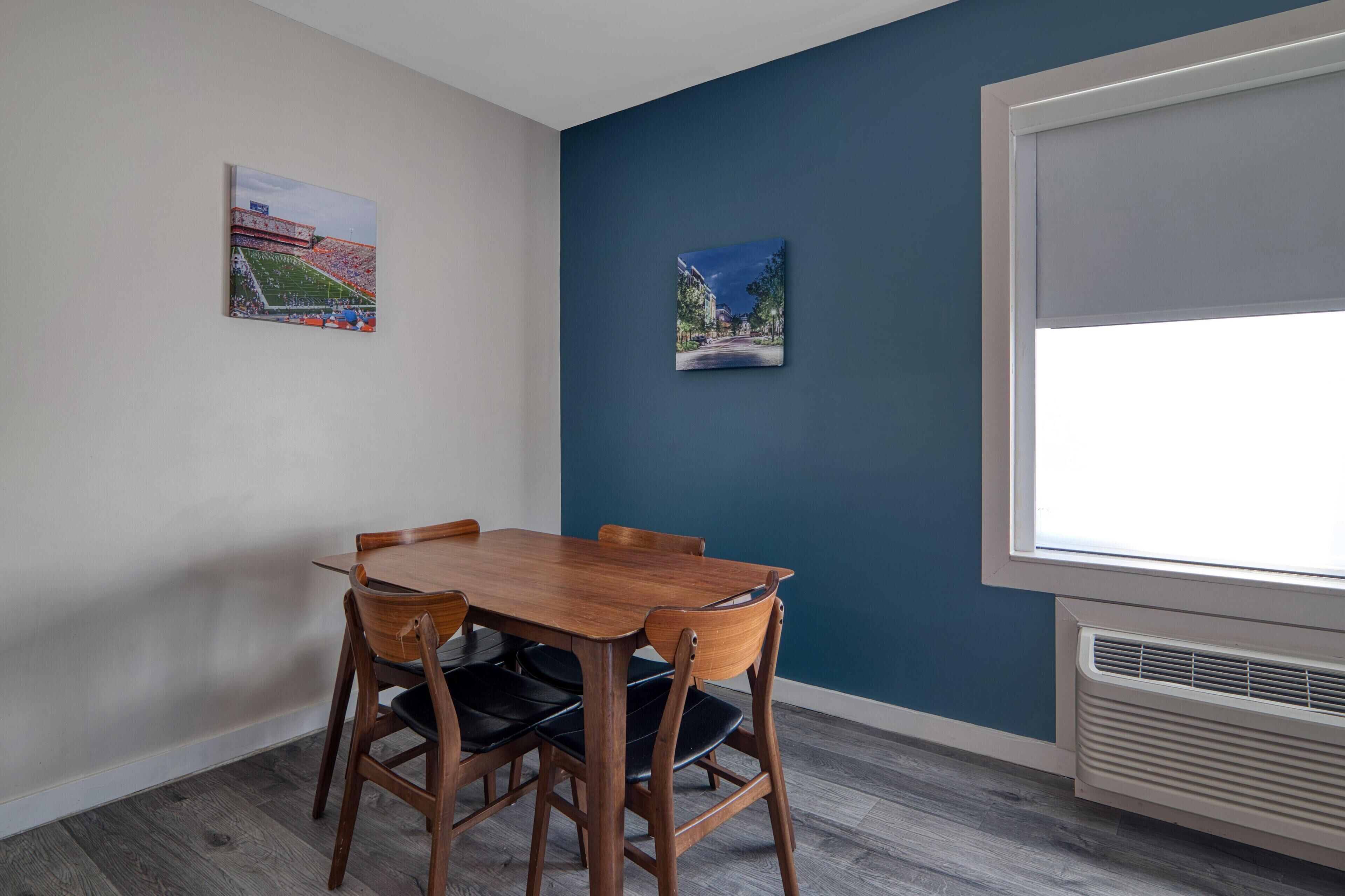 A dining area with a wooden table and chairs, two framed pictures on the wall, and a window with blinds.
