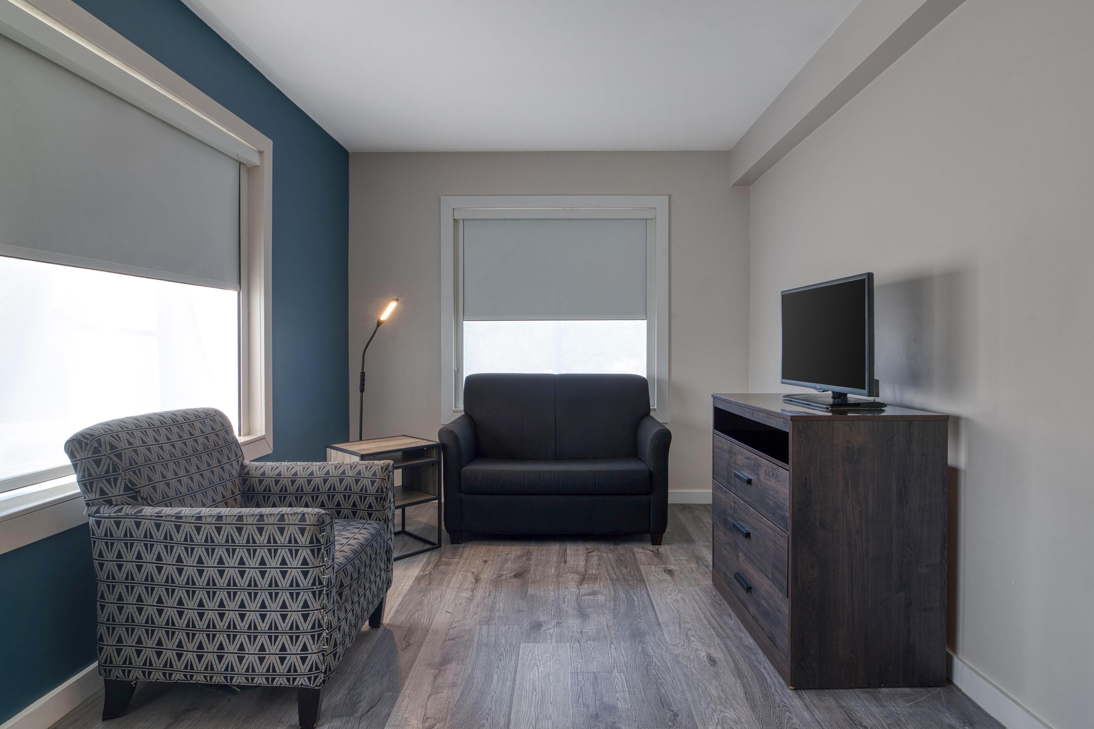 Un petit salon avec un fauteuil à motifs, un canapé foncé, un téléviseur posé sur un meuble en bois et une lampe sur pied.