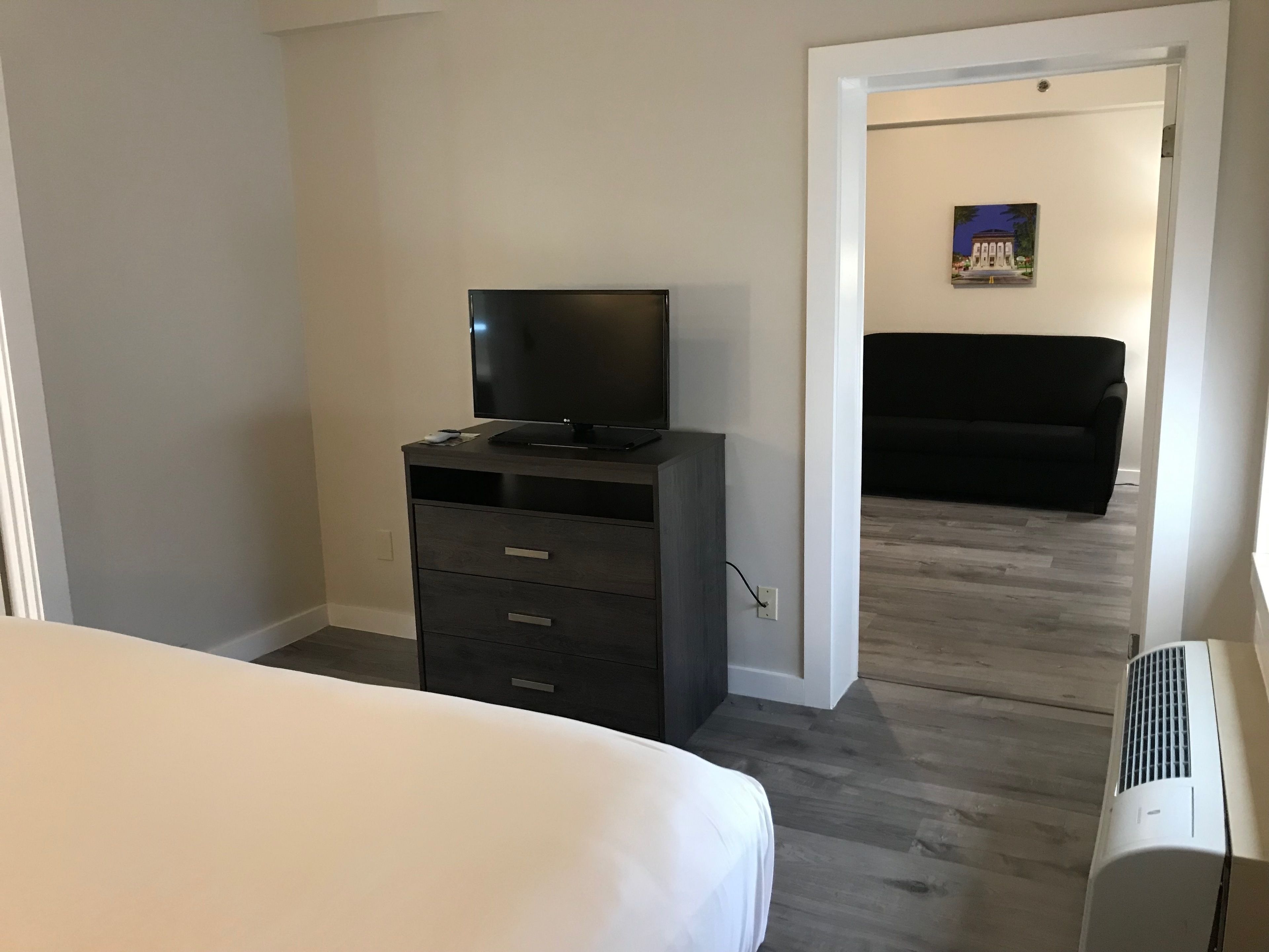 Habitación de hotel con cama, cómoda con televisor, sofá negro y un cuadro en la pared.