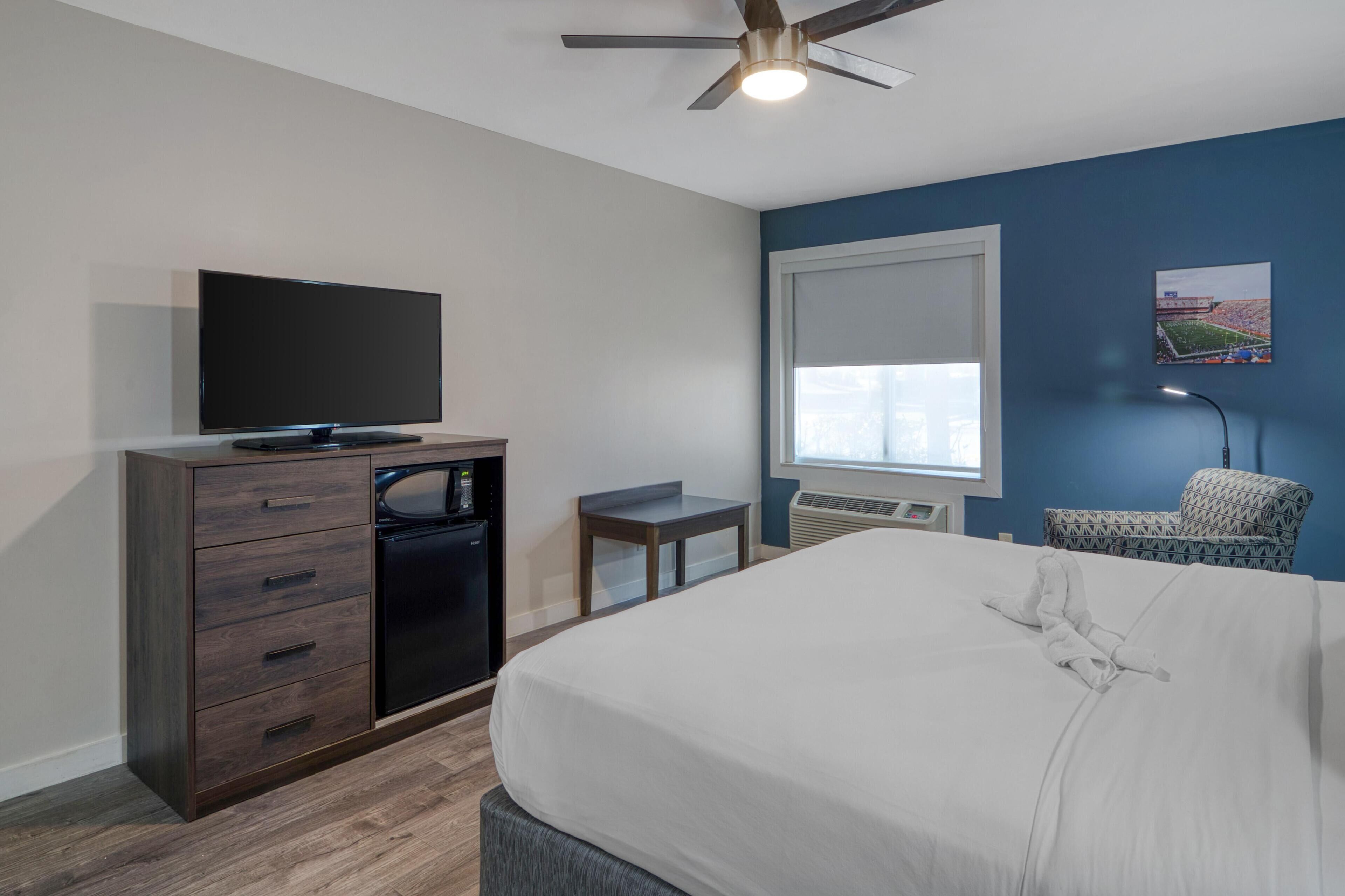 A hotel room with a bed, a TV on a wooden cabinet, a small table, a chair, and a ceiling fan.