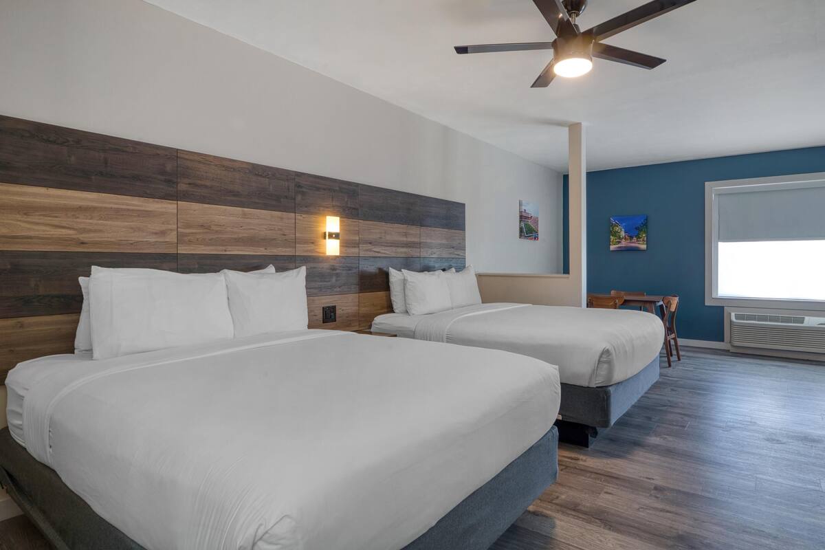 A hotel room with two beds, a ceiling fan, a wall-mounted light, and a window with blinds.
