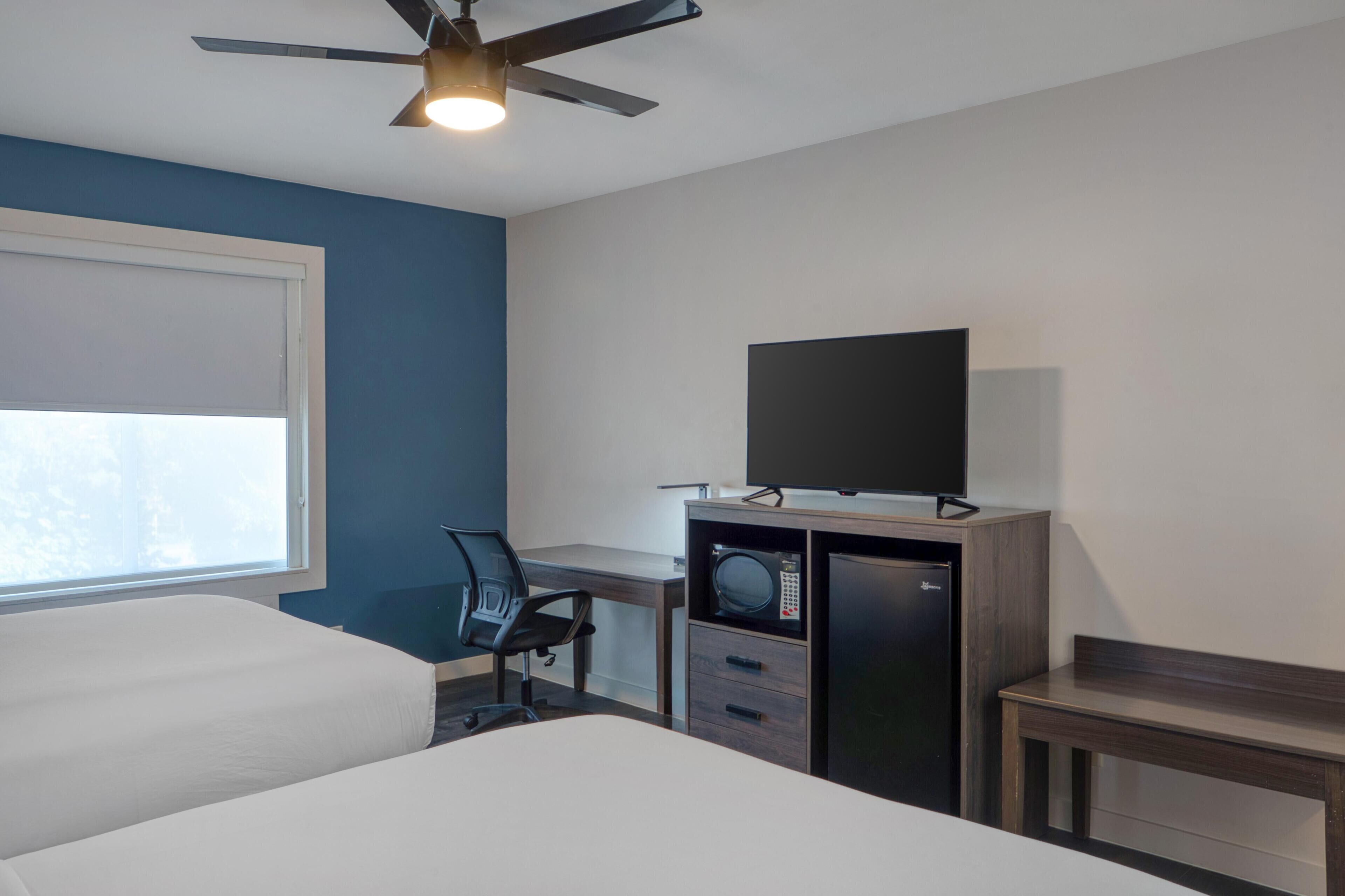 Habitación de hotel con cama, escritorio, silla, televisor, microondas y ventilador de techo.