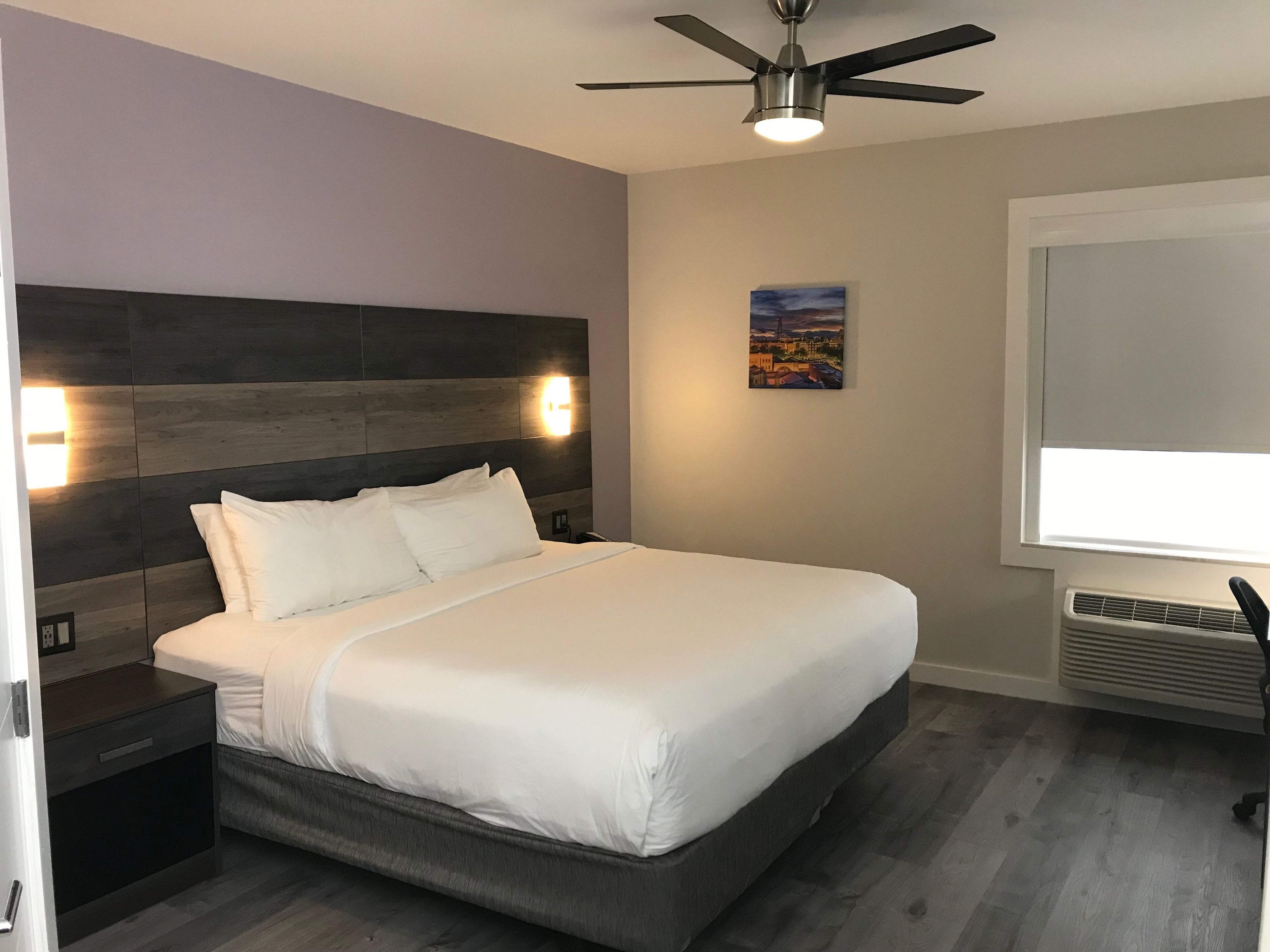 Une chambre moderne avec un grand lit, une tête de lit dotée d’un éclairage intégré, un ventilateur de plafond et un tableau représentant un paysage urbain accroché au mur.