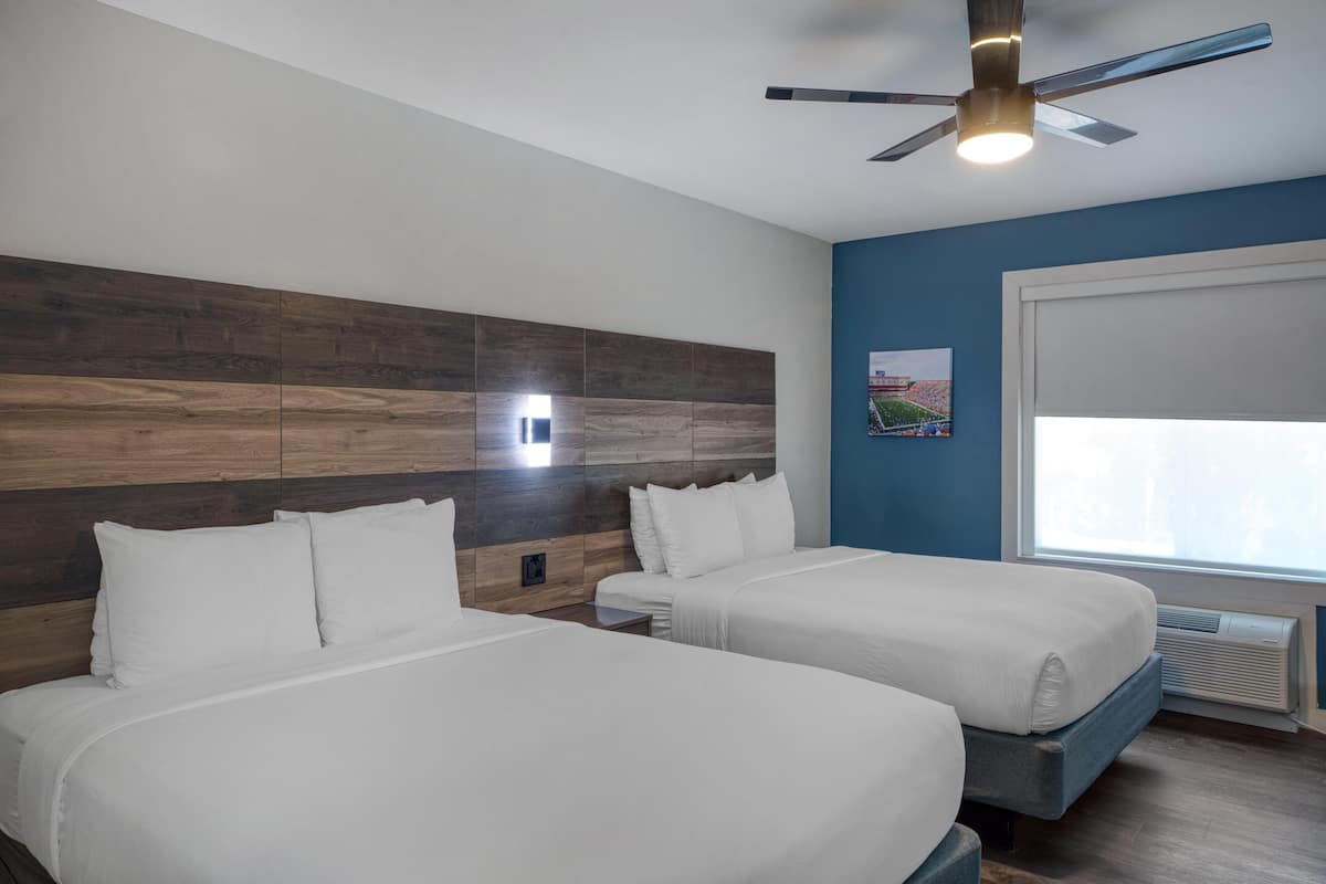 A hotel room with two beds, a ceiling fan, a window with blinds, and a wall-mounted air conditioning unit.