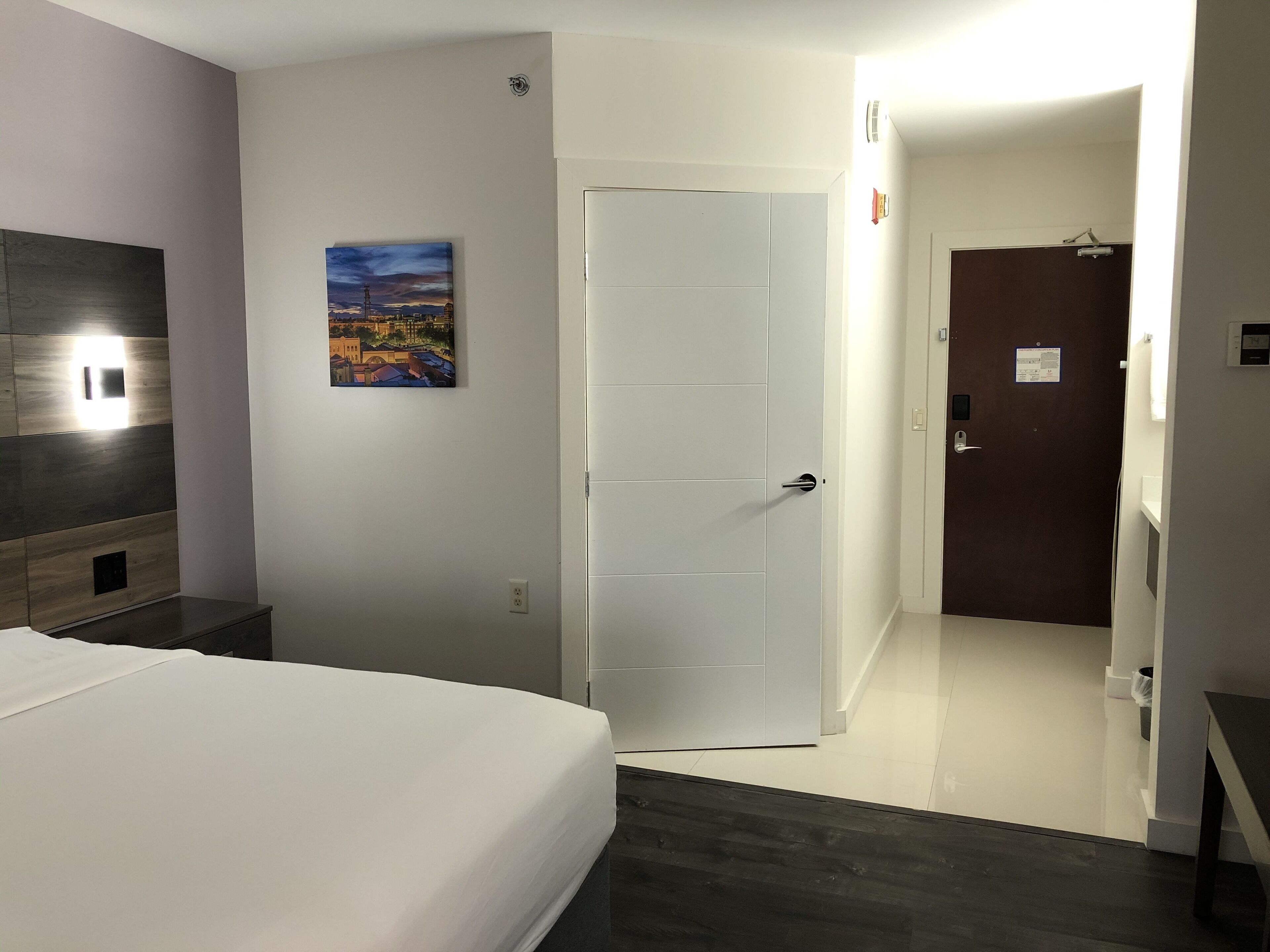 A hotel room with a bed, a door, a painting, and a bathroom visible through an open door.