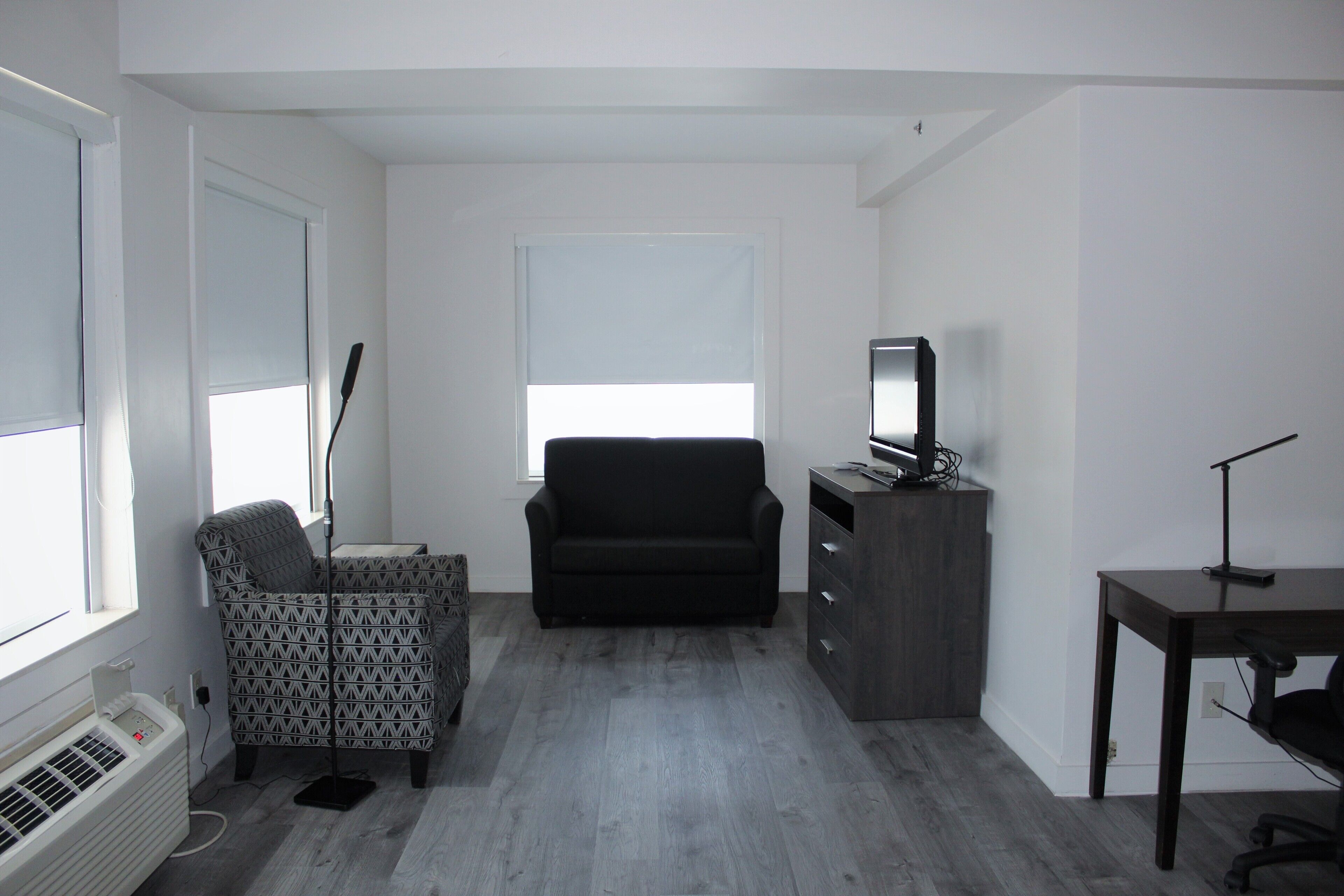 Une pièce de taille modeste, avec un canapé noir, un fauteuil à motifs, un téléviseur posé sur un meuble et un bureau avec une chaise.