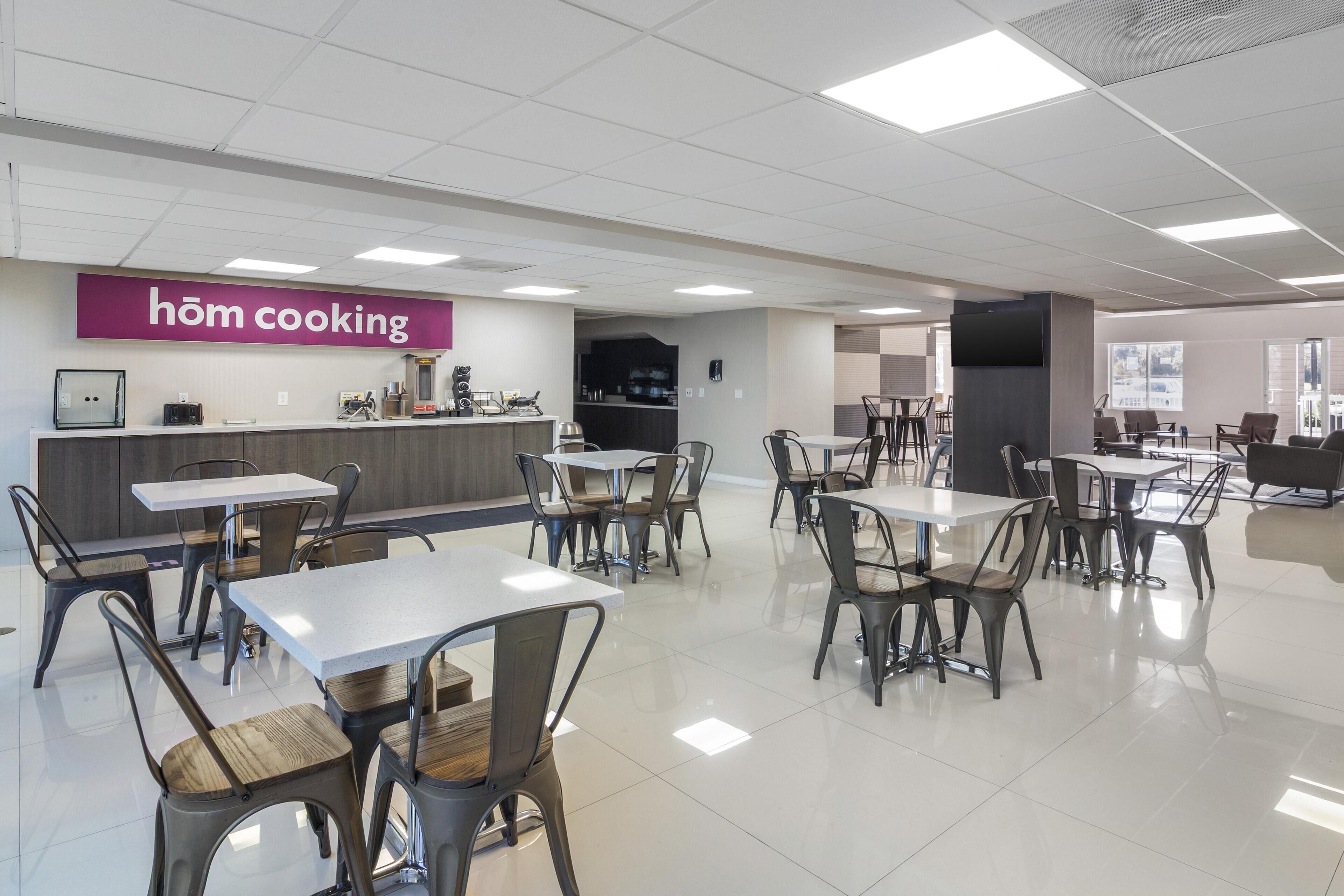 A modern restaurant interior with a 'höm cooking' sign, white tables, and metal chairs.