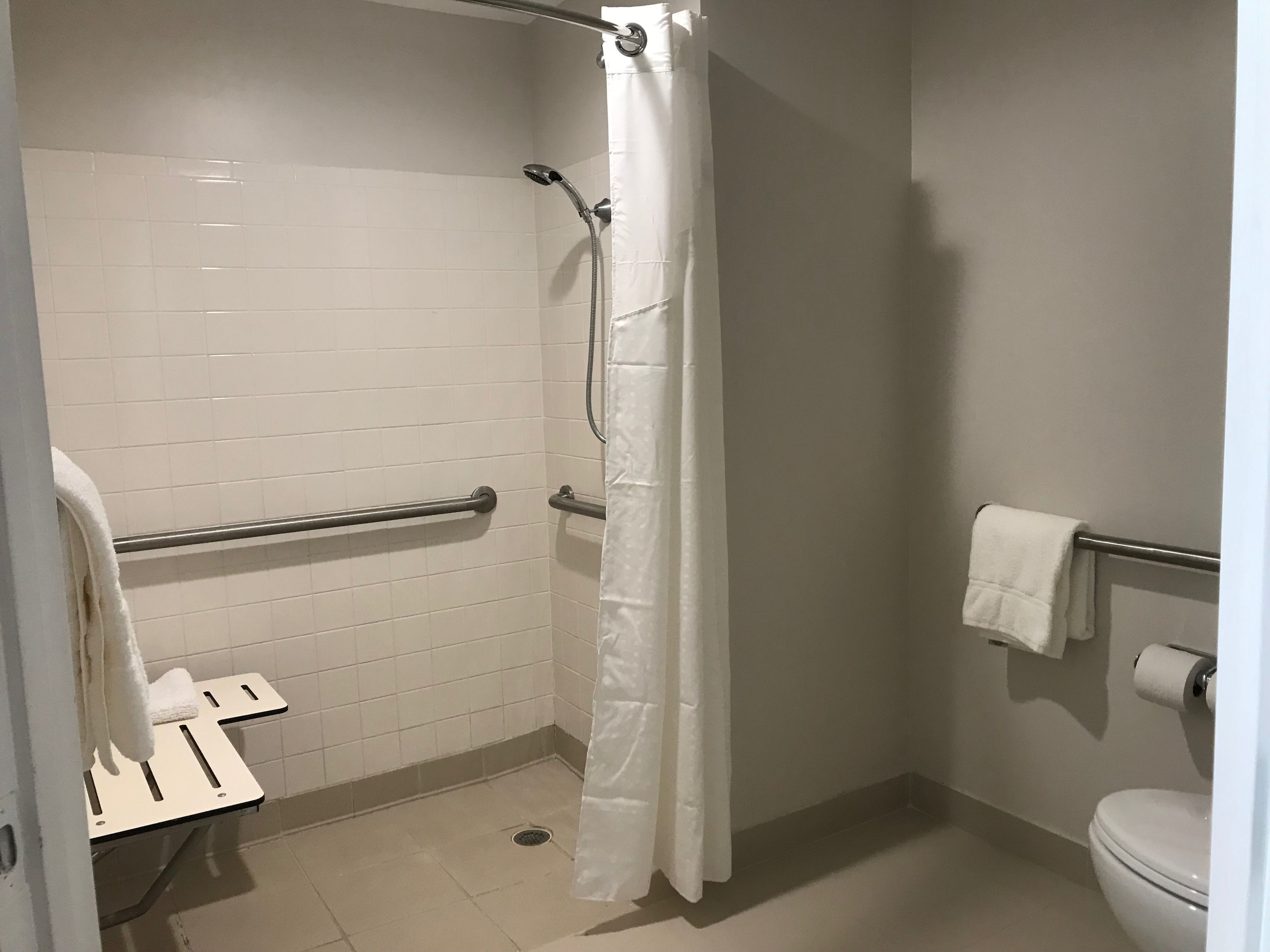 A small, white-tiled bathroom with a shower, a toilet, and a grab bar.