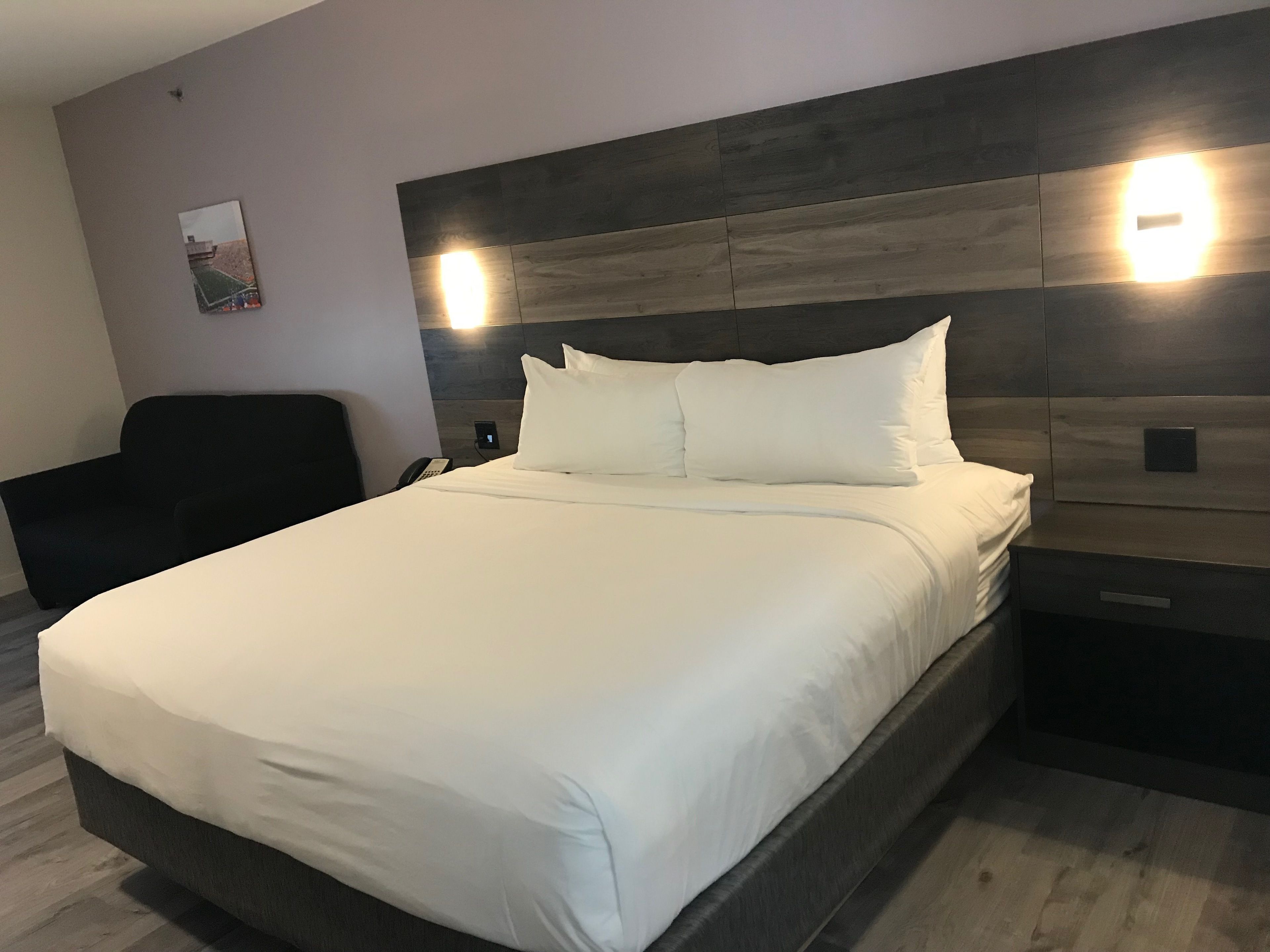 A modern hotel room with a large bed, a nightstand, a chair, and a wall-mounted light fixture.