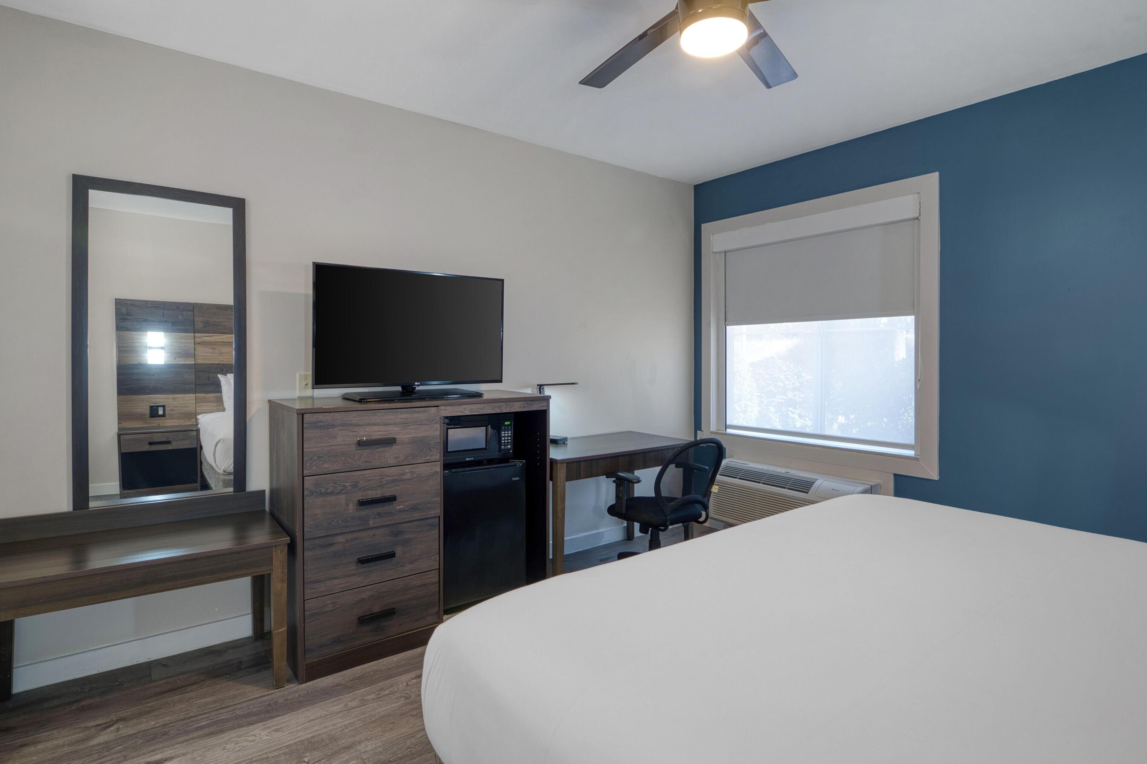 Un dormitorio con cama, escritorio con computadora, silla, cómoda, televisor y ventana con persianas.
