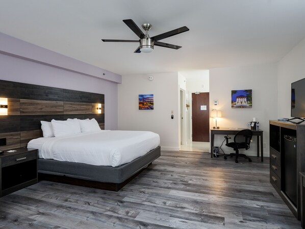 A modern hotel room with a large bed, a desk, a chair, a ceiling fan, and a television.