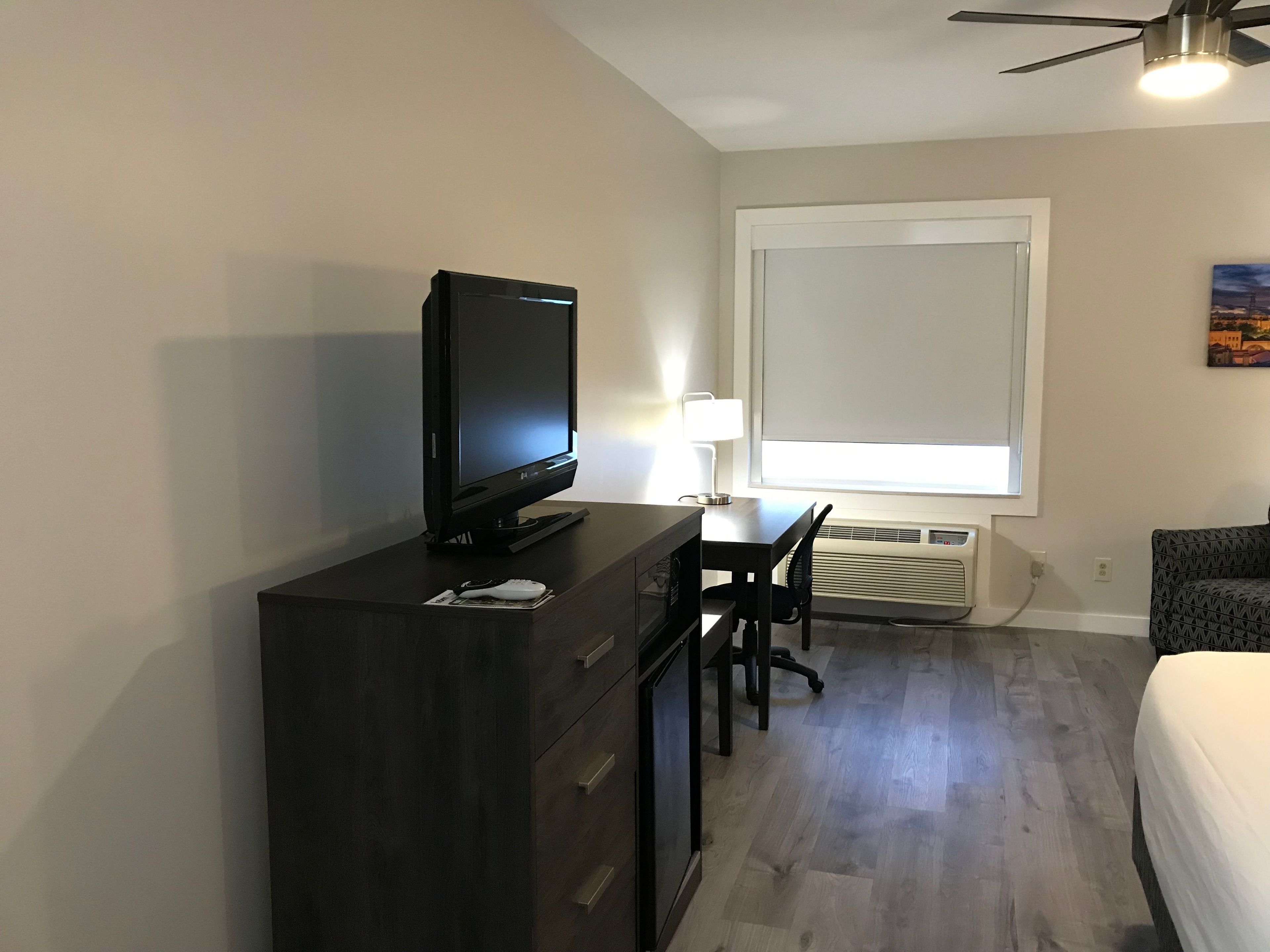Habitación de hotel con cómoda de madera oscura, televisor de pantalla plana, escritorio con silla, ventana con persianas, ventilador de techo y cama.
