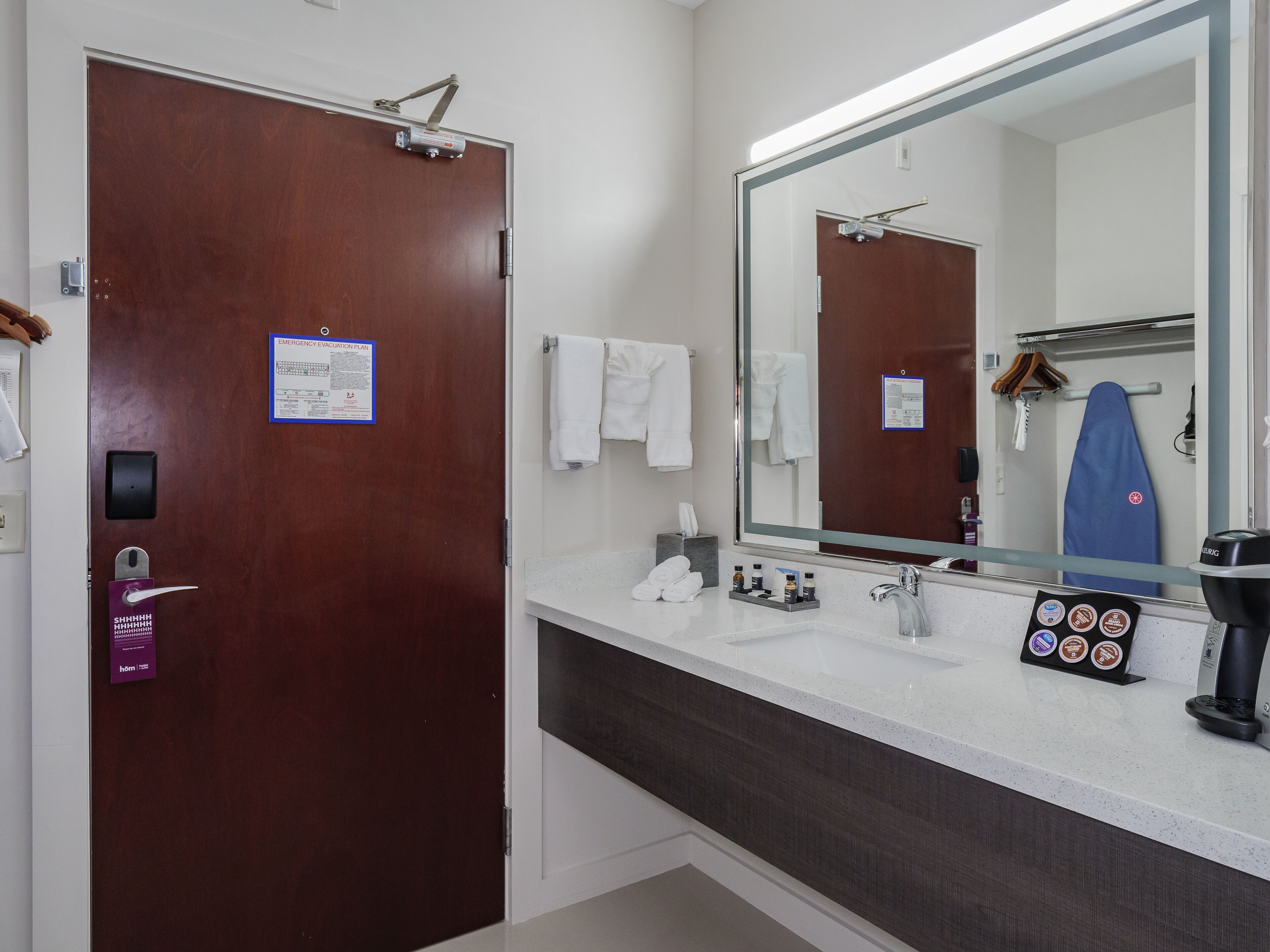 A hotel bathroom with a wooden door, a large mirror, a sink, and a coffee machine.