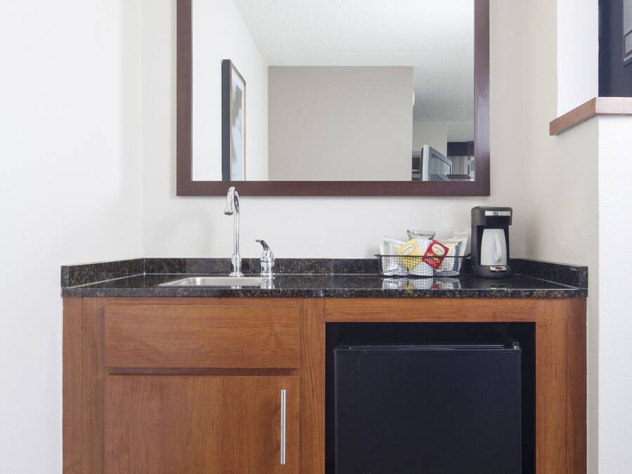 Un baño de hotel pequeño y funcional con lavamanos, espejo y un refrigerador compacto.