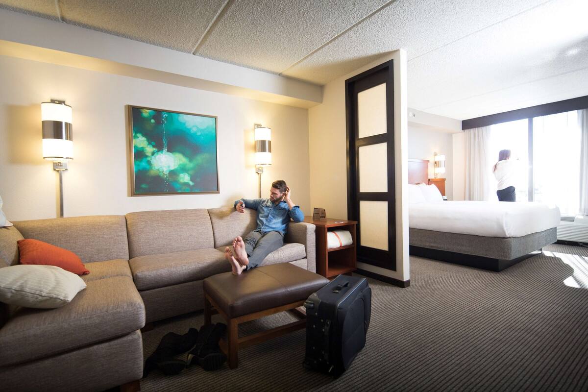 A hotel room with a sofa, a bedside table, a lamp, a painting, and a bed.
