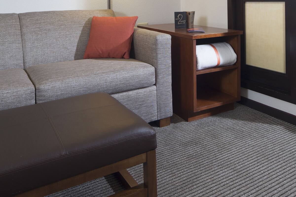 A hotel room with a gray sofa, a red pillow, a wooden bench, a wall-mounted lamp, a wooden nightstand, and a large window with a view of the sky.