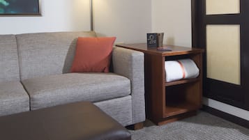Habitación de hotel con sofá gris, almohada roja, banco de madera, lámpara de pared, mesita de noche de madera y un ventanal con vistas al cielo.