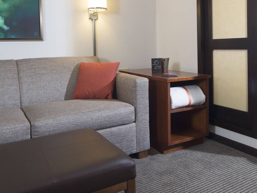 Habitación de hotel con sofá gris, almohada roja, banco de madera, lámpara de pared, mesita de noche de madera y un ventanal con vistas al cielo.