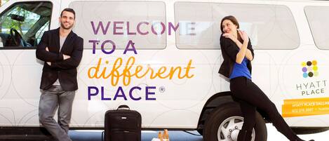 A white van with "WELCOME TO A DIFFERENT PLACE" and "HYATT PLACE" branding, luggage, and a person leaning on the van.