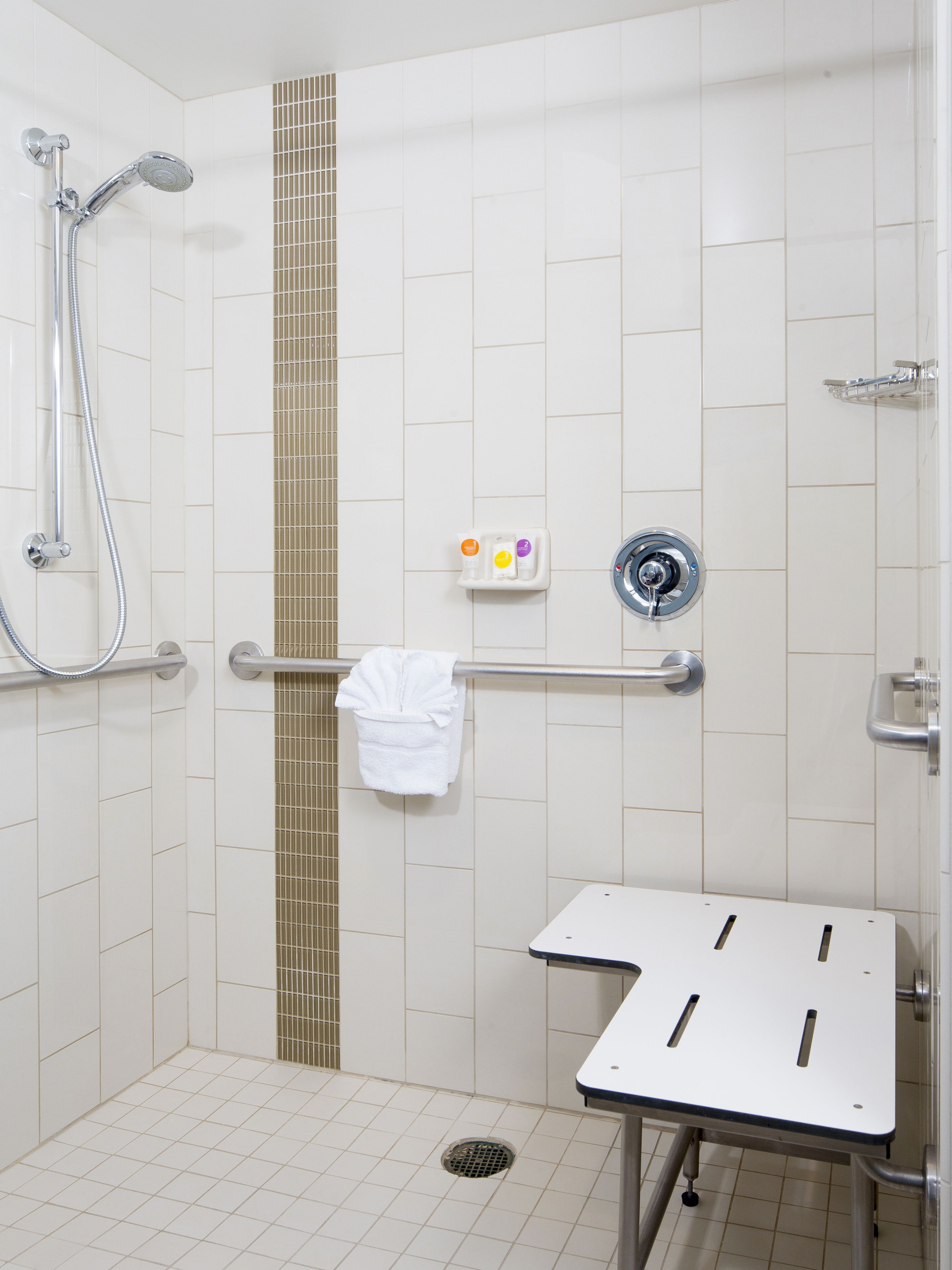 Une salle de bain avec des carreaux blancs, une douche, une barre d’appui, un porte-serviettes et des toilettes.