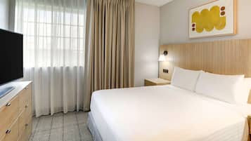 Quarto de hotel com cama, televisão, mesa de cabeceira, luminária e janela com cortinas.