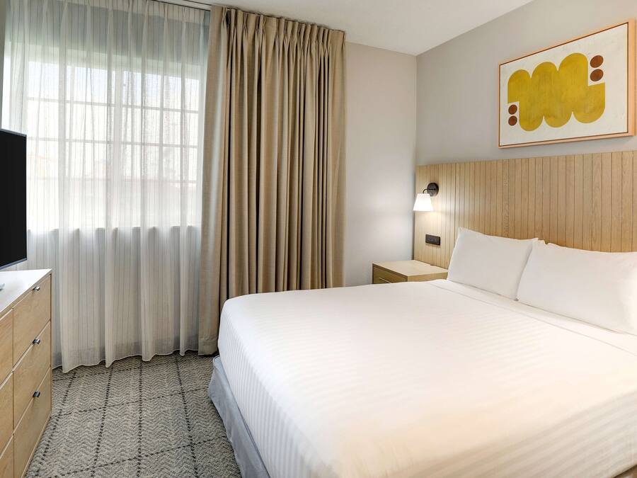 Quarto de hotel com cama, televisão, mesa de cabeceira, luminária e janela com cortinas.