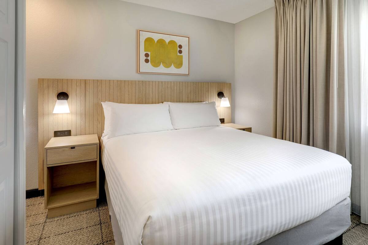 A hotel room with a large bed, two bedside tables with lamps, a wooden headboard, and a wall-mounted artwork.