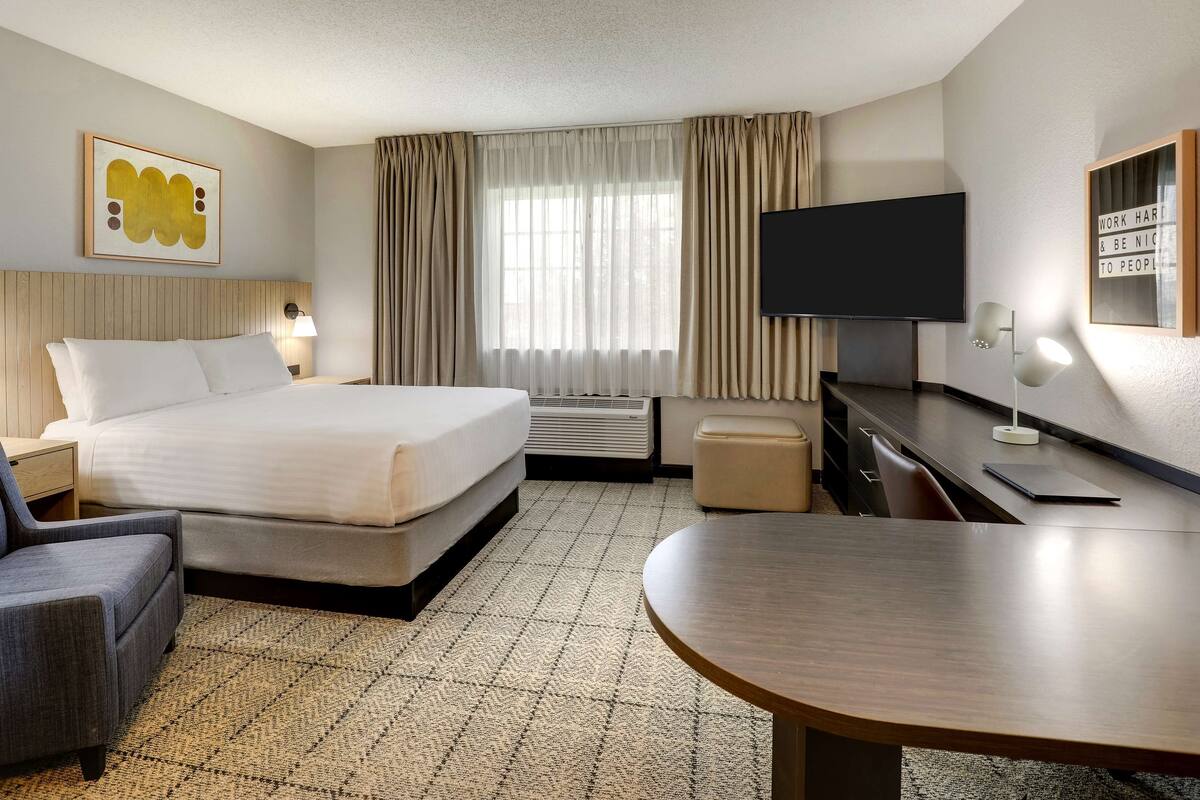 A hotel room with a bed, a desk, a chair, a TV, and a large window with curtains.